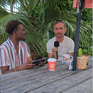 Two people sitting at a wooden table outdoors, one holding a tablet and the other holding a microphone, with tropical plants in the background.