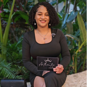 A woman with curly hair smiling while sitting outdoors among green plants, holding a black sign that reads 'Authentic'.