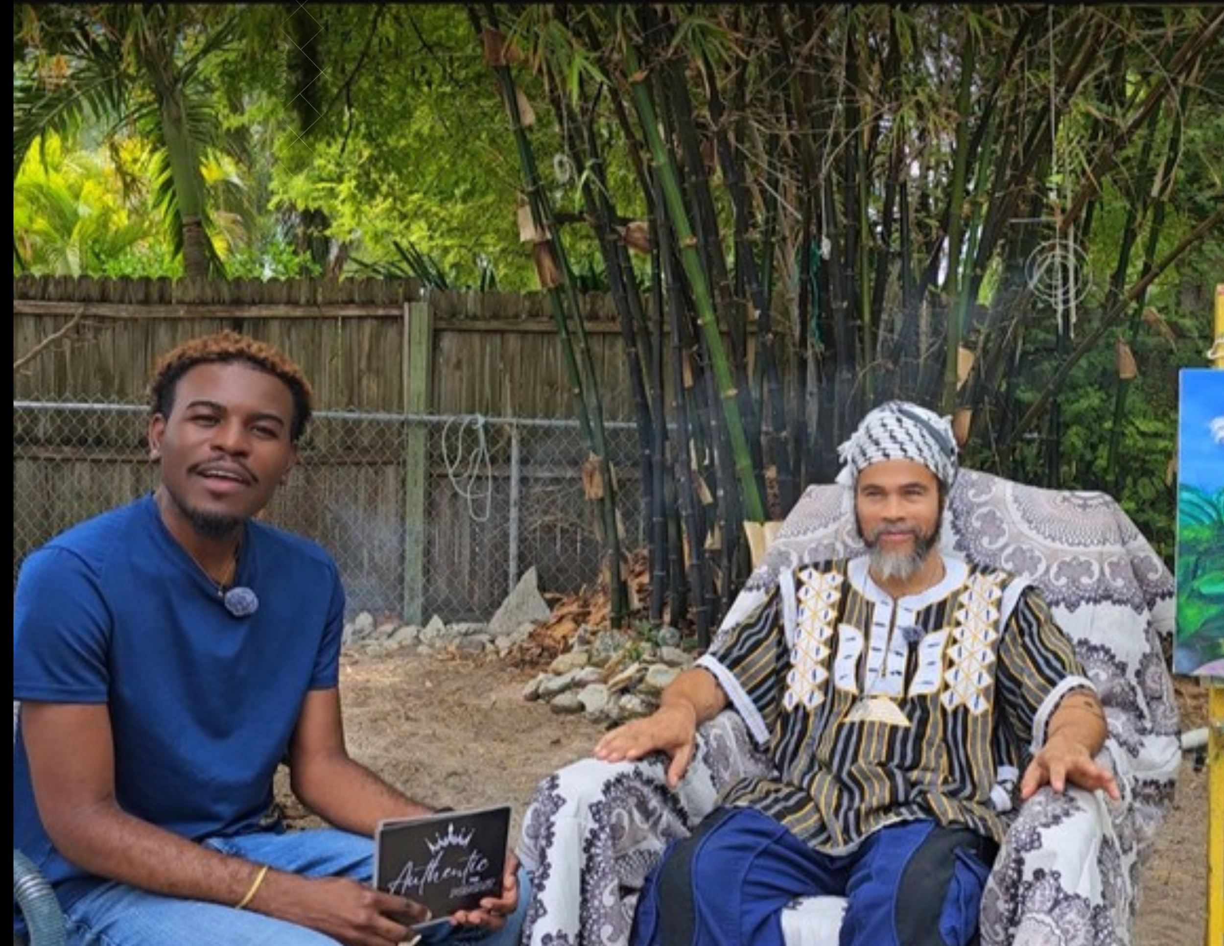 Two men are sitting outdoors in a backyard with a wooden fence and green trees in the background. One man is younger with dark skin, brown hair, wearing a blue shirt, and holding a small black chalkboard sign. The other man is older with light skin, 