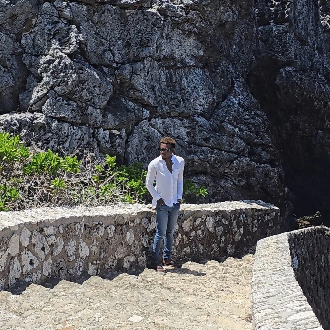 A man in a white shirt and jeans stands on a stone pathway next to green bushes and a large rock formation in the background.