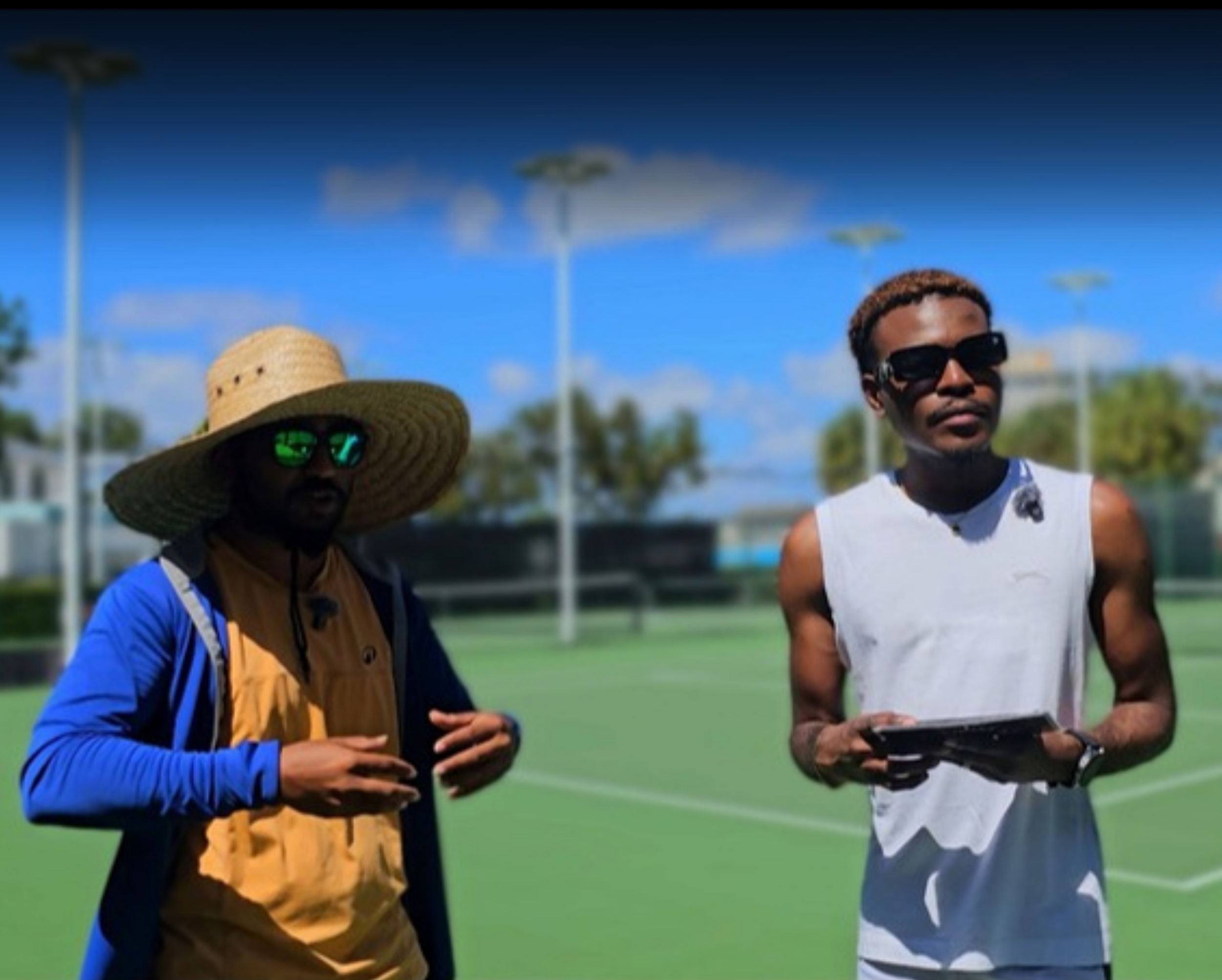 Two men are outdoors on a sports field. The man on the left is wearing a large straw hat, dark sunglasses, a blue jacket, and a yellow vest, holding a device in his hands. The man on the right has curly hair, wearing sunglasses, a white sleeveless sh