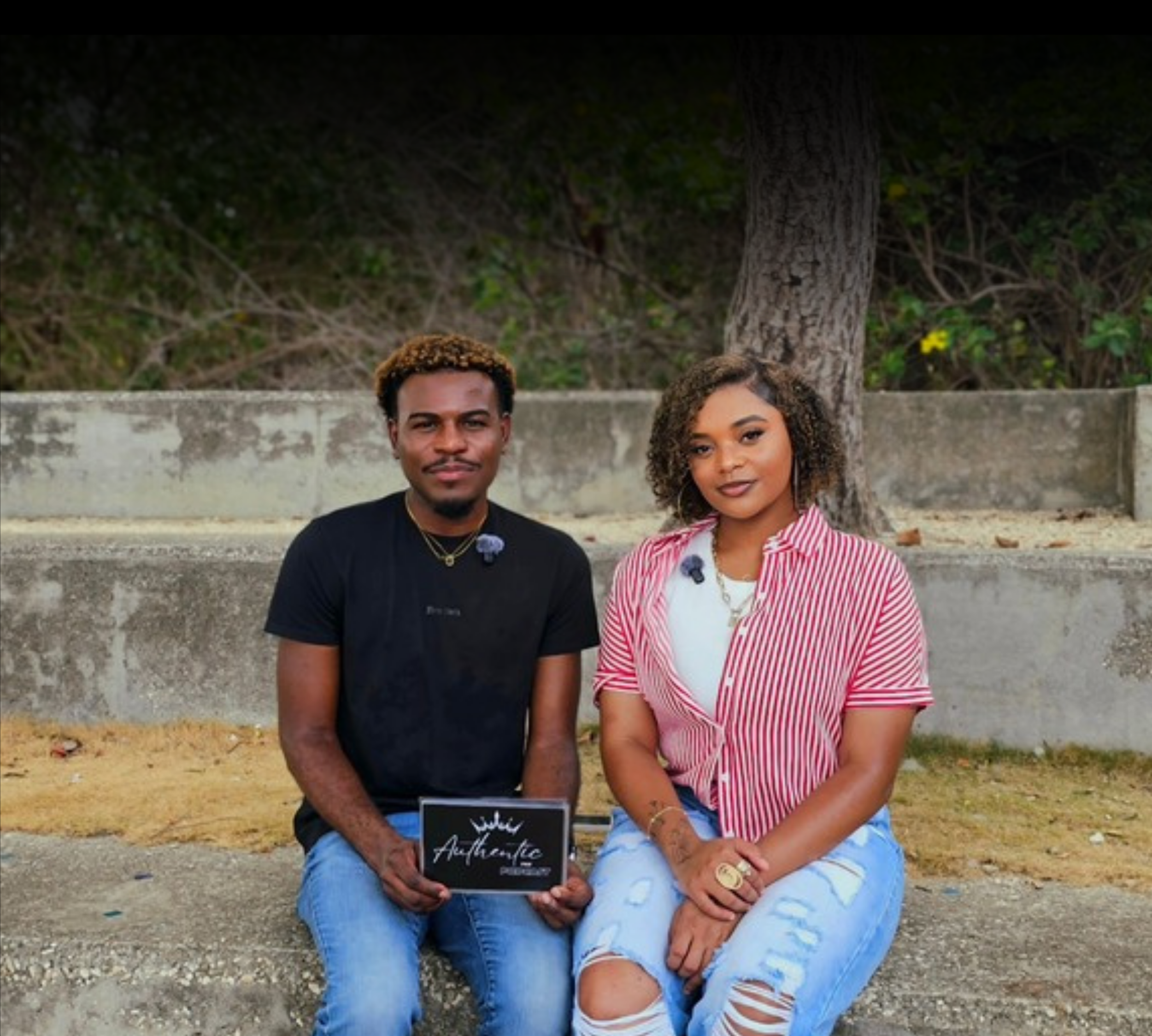 A young man and woman sitting outdoors under a tree, wearing casual clothing and smiling. The man is holding a black sign with white text that reads "Authentic."