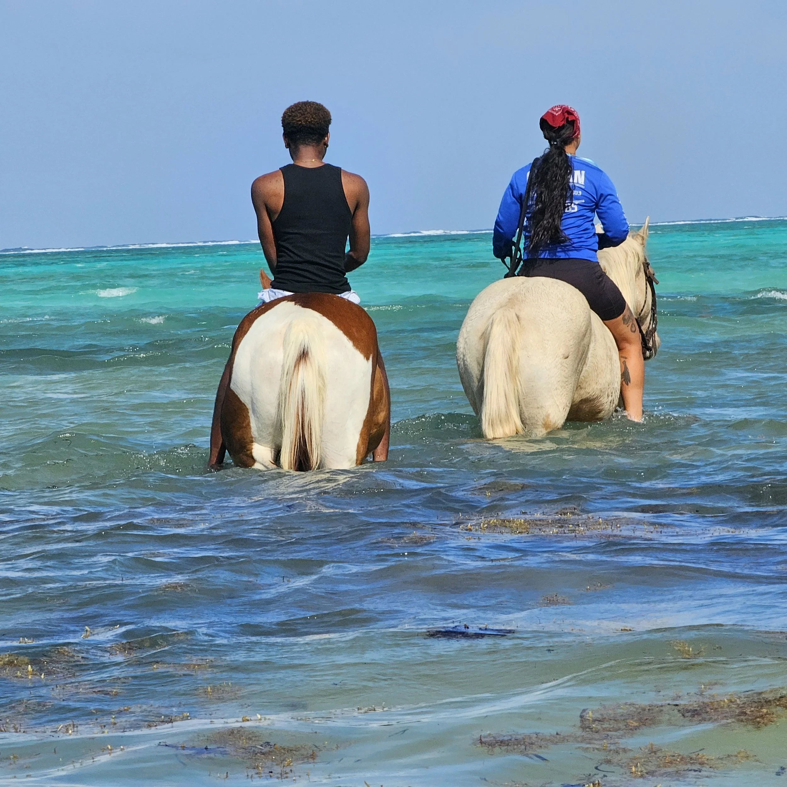 Two women riding horses in shallow ocean water with waves and a clear blue sky in the background.