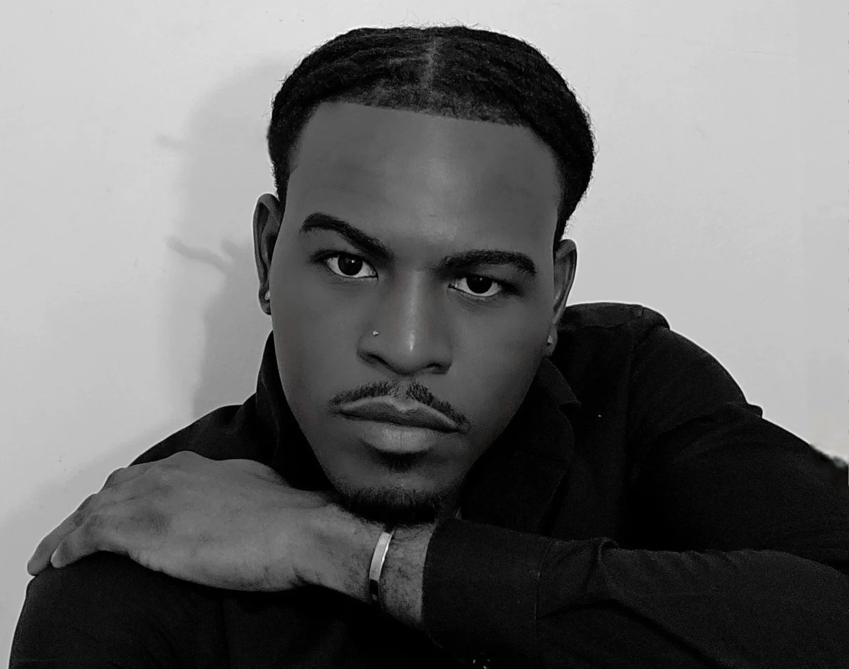Black and white close-up portrait of a young man with short hair, a mustache, and a small nose piercing, wearing a dark shirt, looking directly at the camera with a serious expression, against a plain light-colored background.