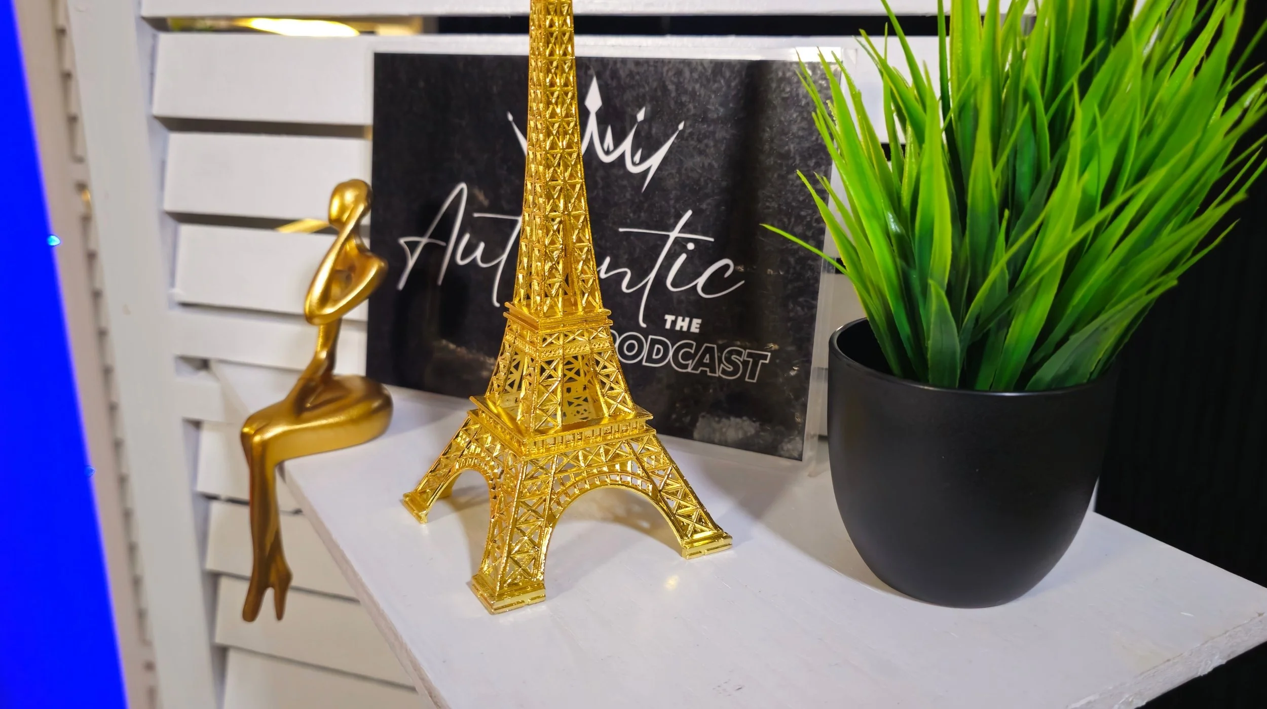 Decorative gold Eiffel Tower model, gold rocking horse, framed black and white sign reading 'Authentic the Podcast,' and a potted green plant on a white shelf.