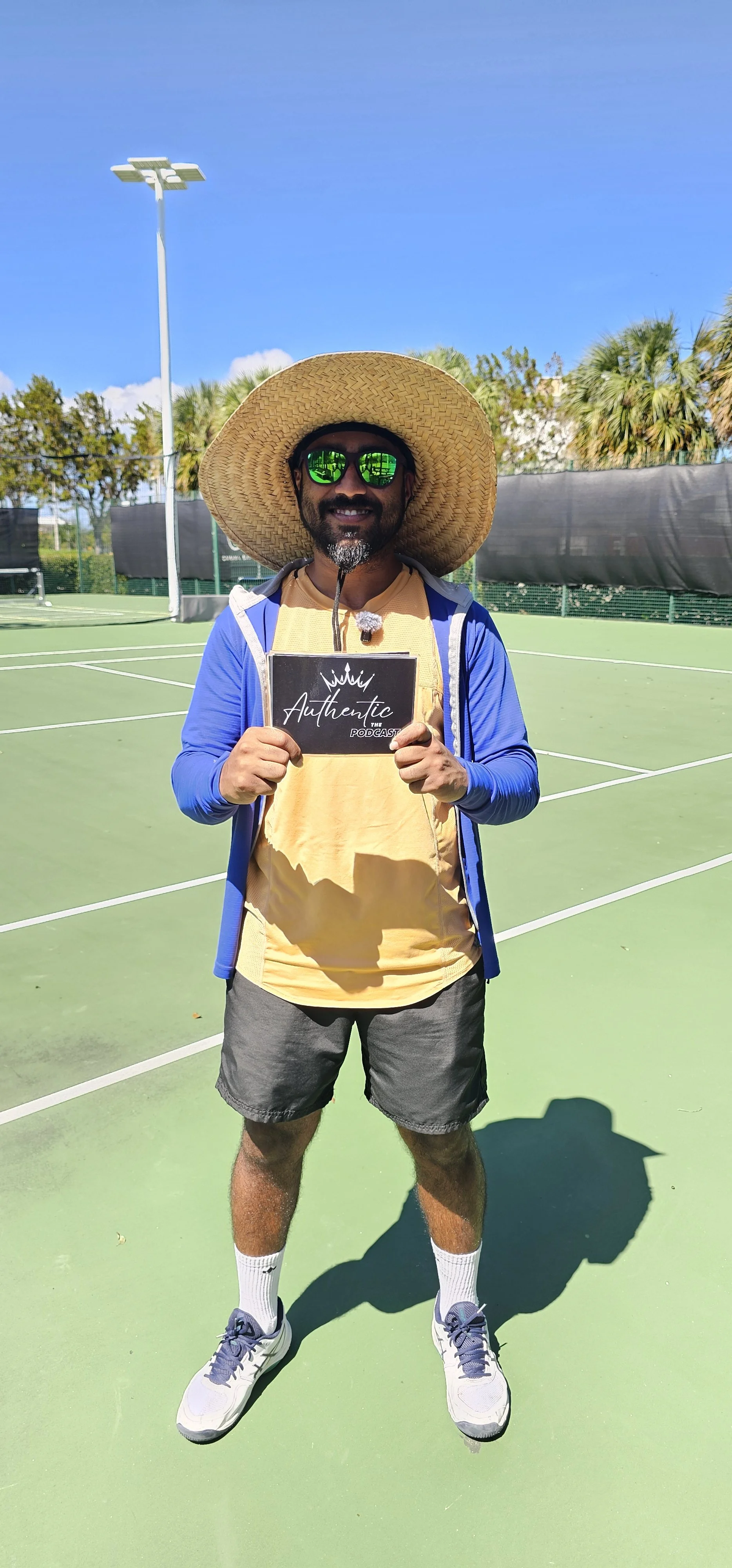 Man in sunglasses and a large sunhat holding a sign that says "Authentic Podcast" on a tennis court under a clear blue sky.