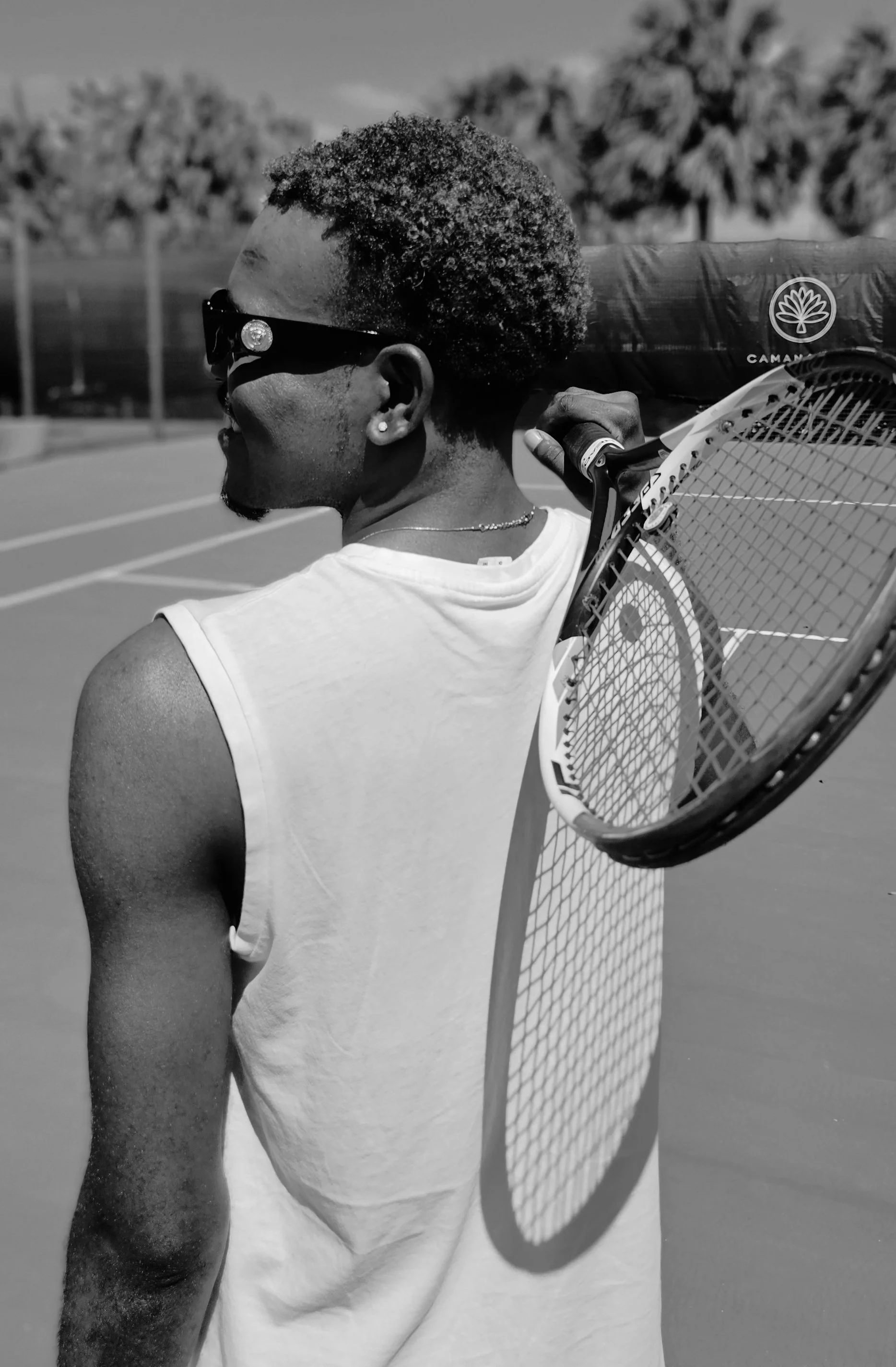 Black and white photo of a woman holding a tennis racket on her shoulder, wearing sunglasses and a sleeveless shirt, outdoors with a tennis court and trees in the background.