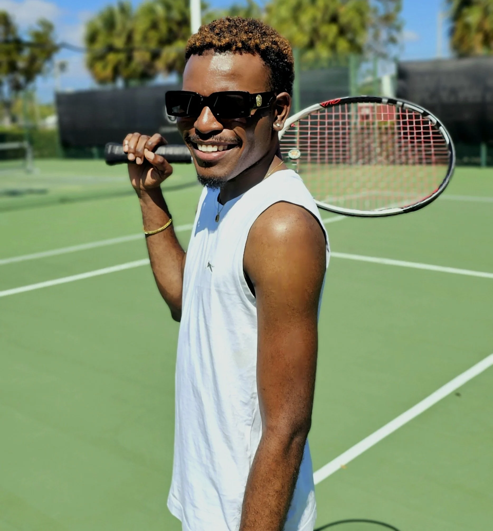 A smiling young man with dark skin and short, curly hair is holding a tennis racket on an outdoor tennis court. He is wearing sunglasses, a white sleeveless shirt, and a gold necklace. The background shows trees, a blue sky, and part of the tennis co