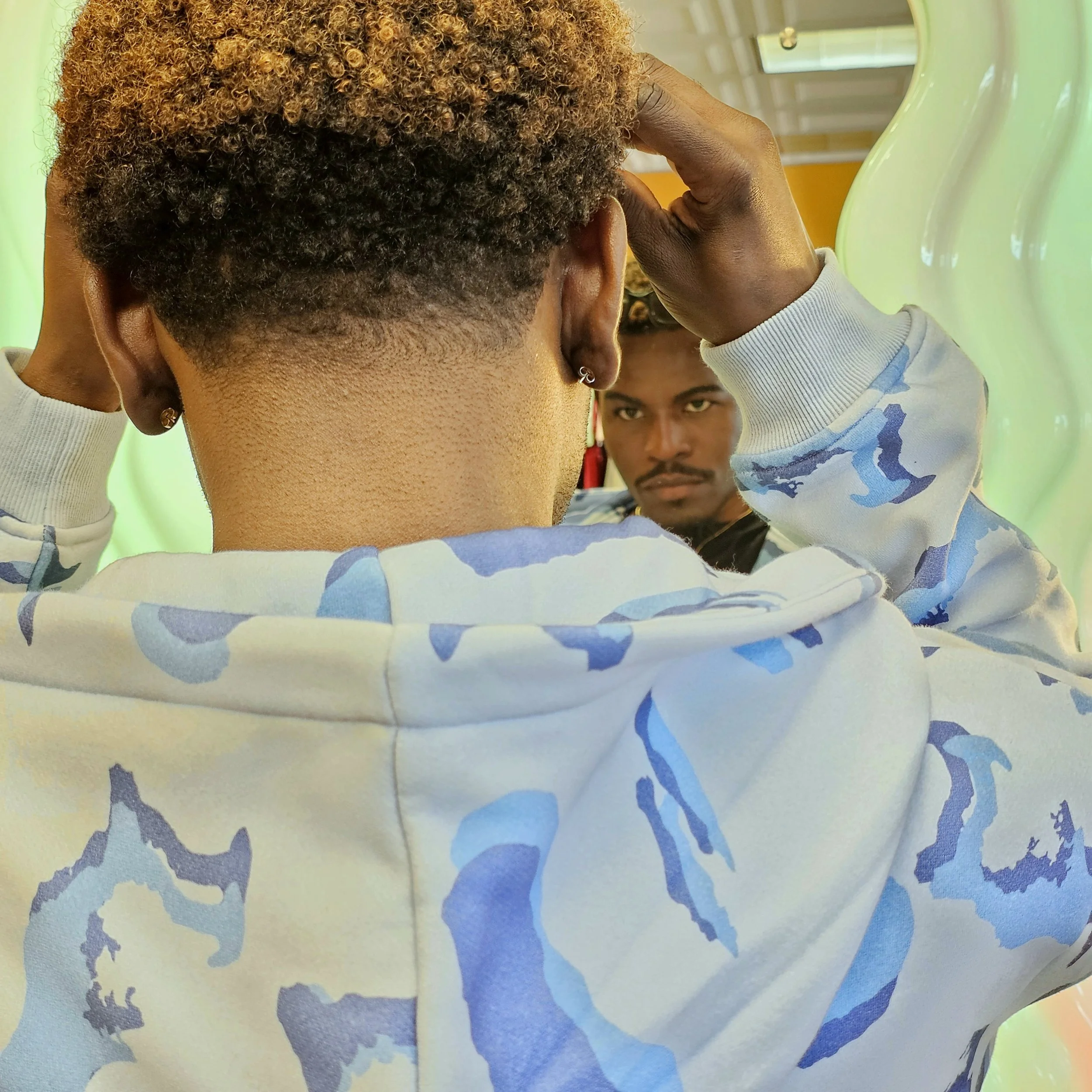 A person with curly hair wearing a light-colored hoodie, looking at a mirror, with another person in the background observing.