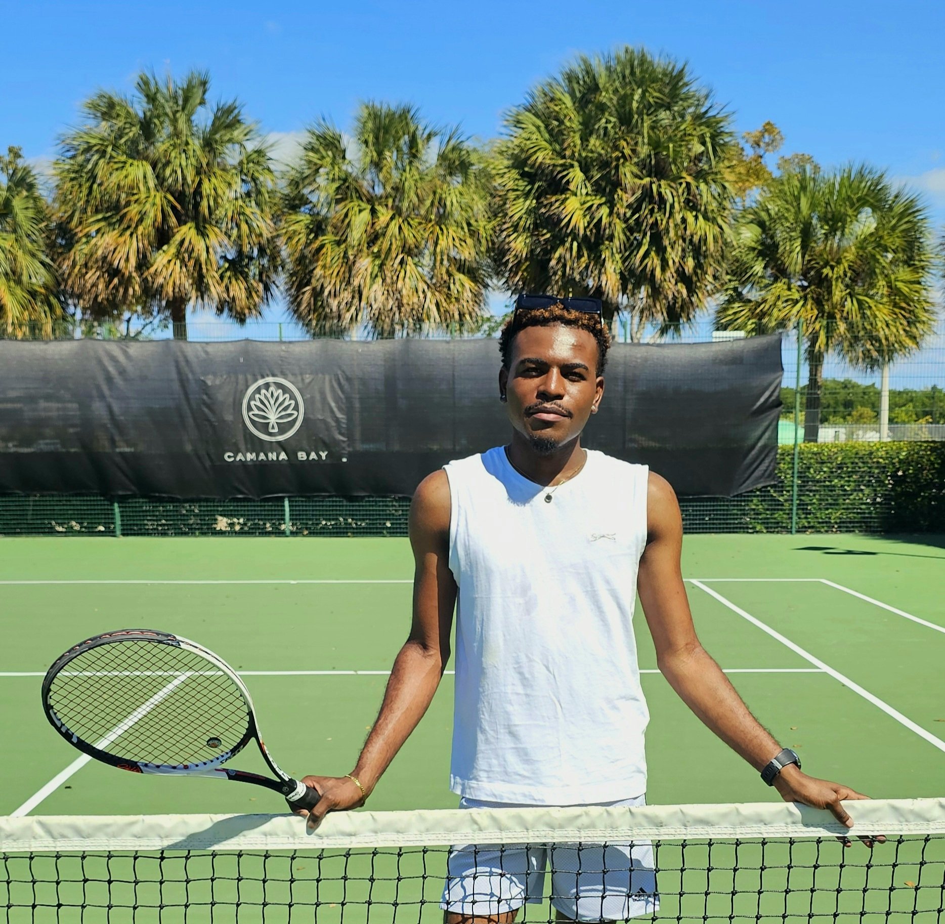 A man with short curly hair and a goatee holding a tennis racket on a tennis court, with palm trees and a blue sky in the background.