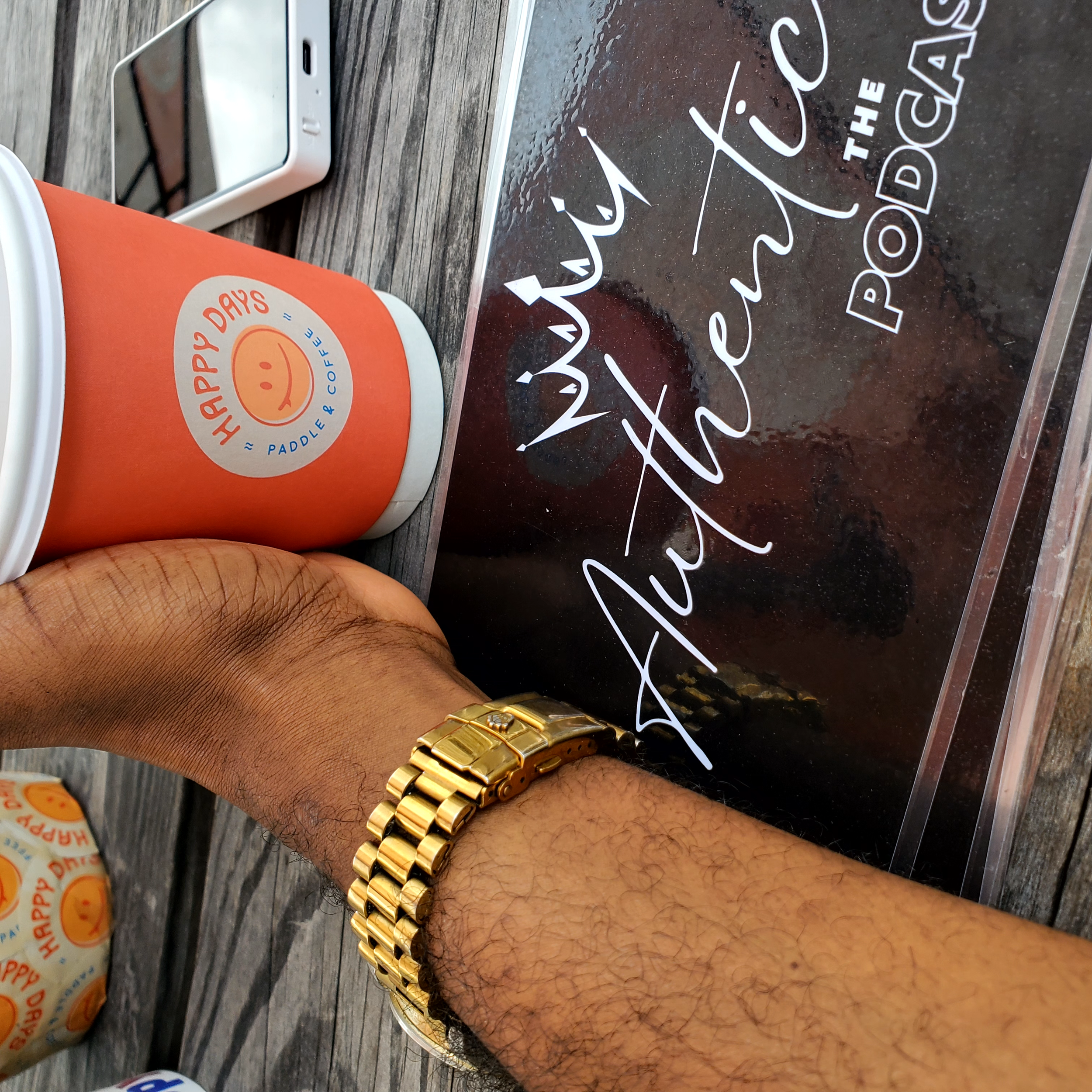 A person holding a paper cup with an orange sleeve that has a smiling face logo and the words "Happy Days" and "Paddle & Coffee" printed on it. The person has a gold watch on their wrist and is sitting at a wooden table next to a smartphone and some 