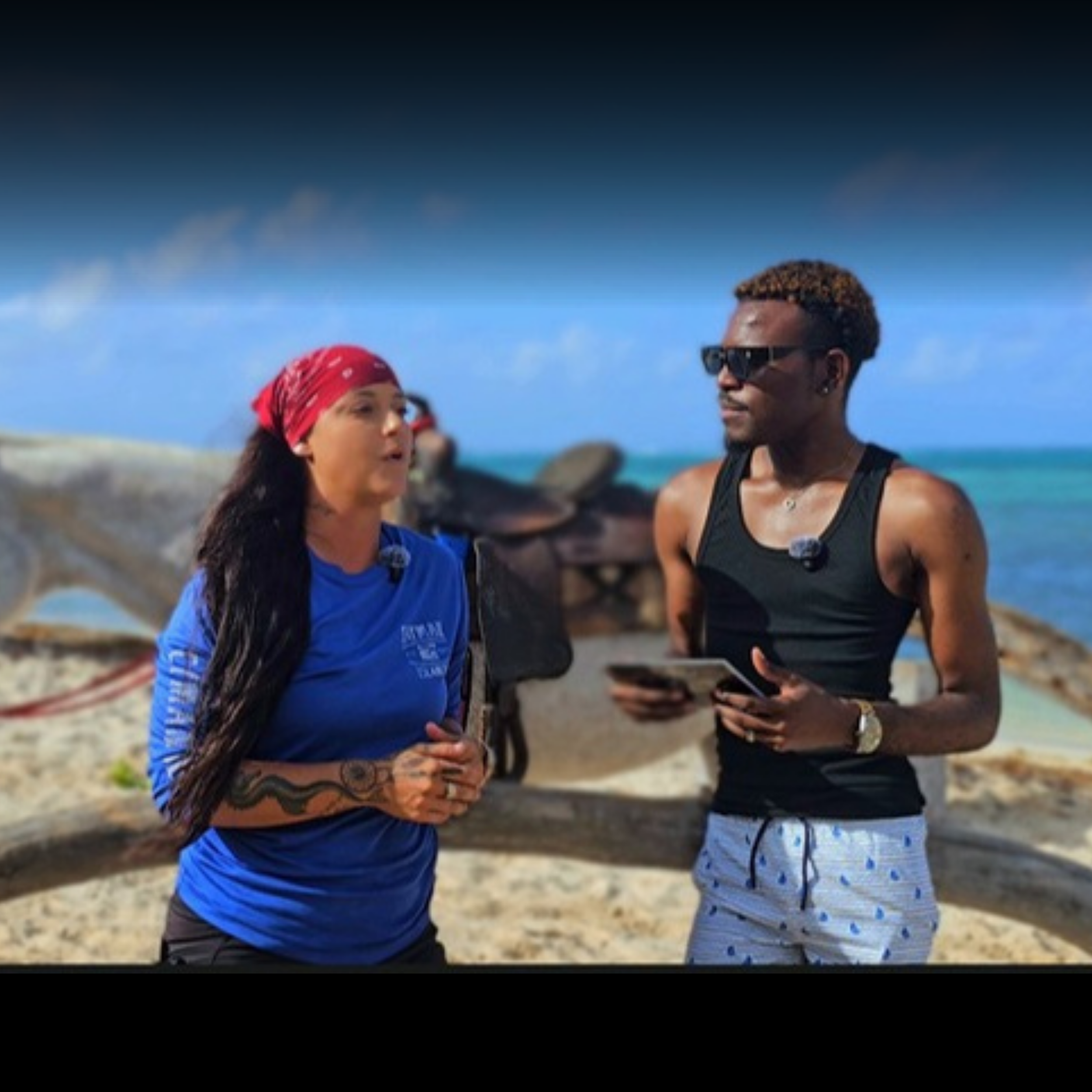 A woman with long dark hair wearing a red bandana and blue shirt, and a man with short curly hair, sunglasses, and a black tank top, talking to each other outdoors under a clear blue sky.