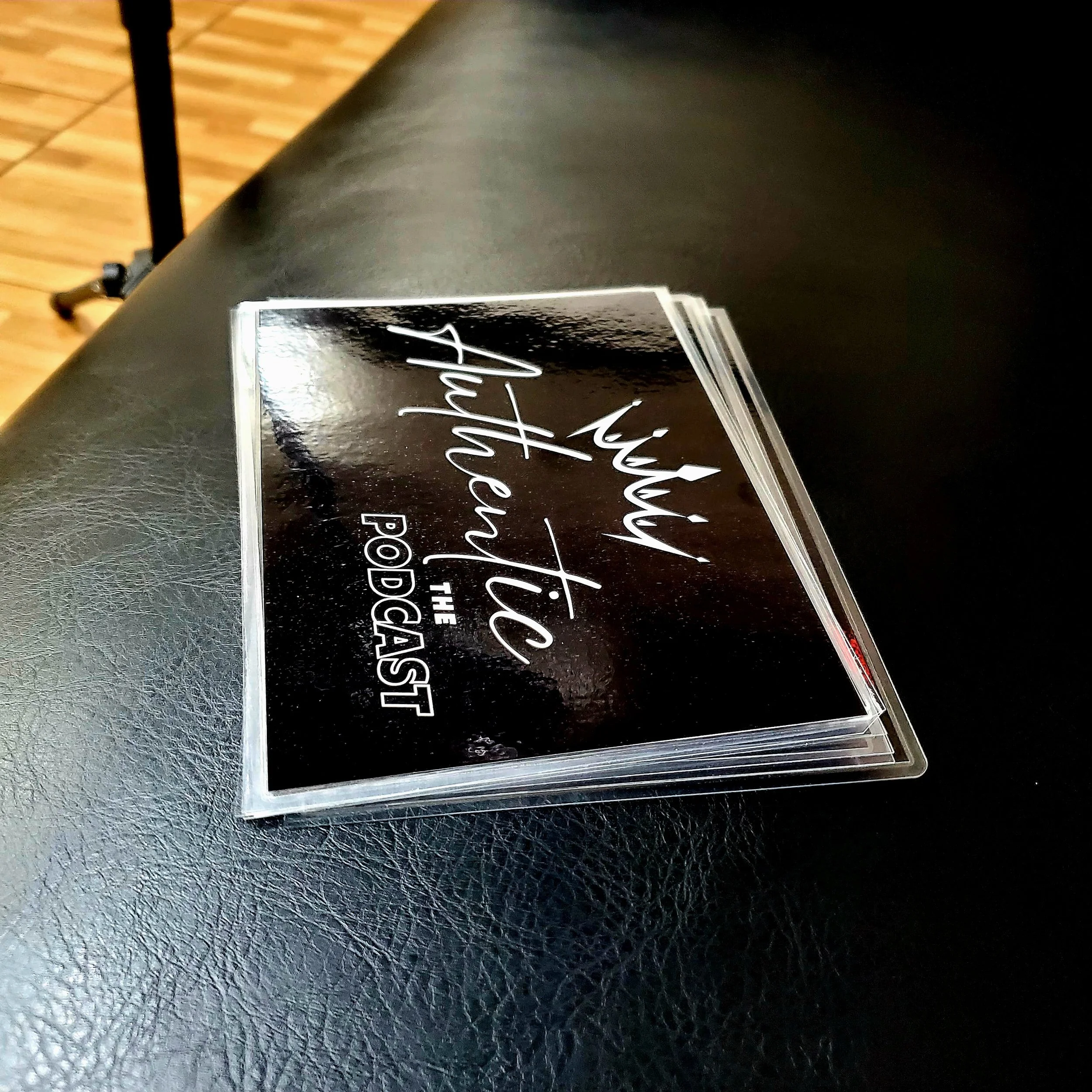 Close-up of a promotional sticker with white cursive writing on a black background, placed on a dark surface. Part of a black table and a wooden floor are visible nearby.