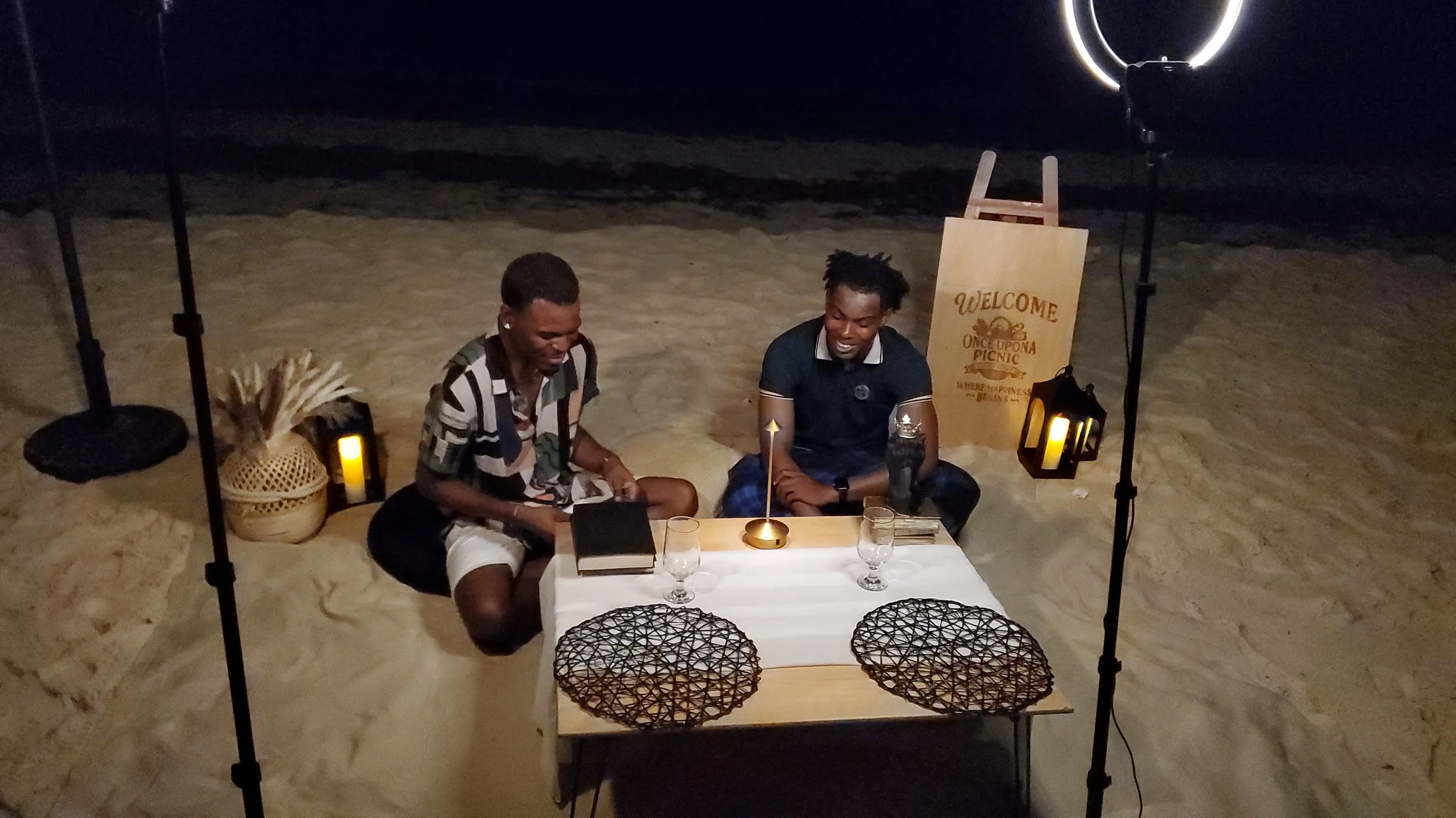 Two men sitting at a small table on the beach at night, with a welcome sign and lanterns around them, enjoying a meal or drinks.
