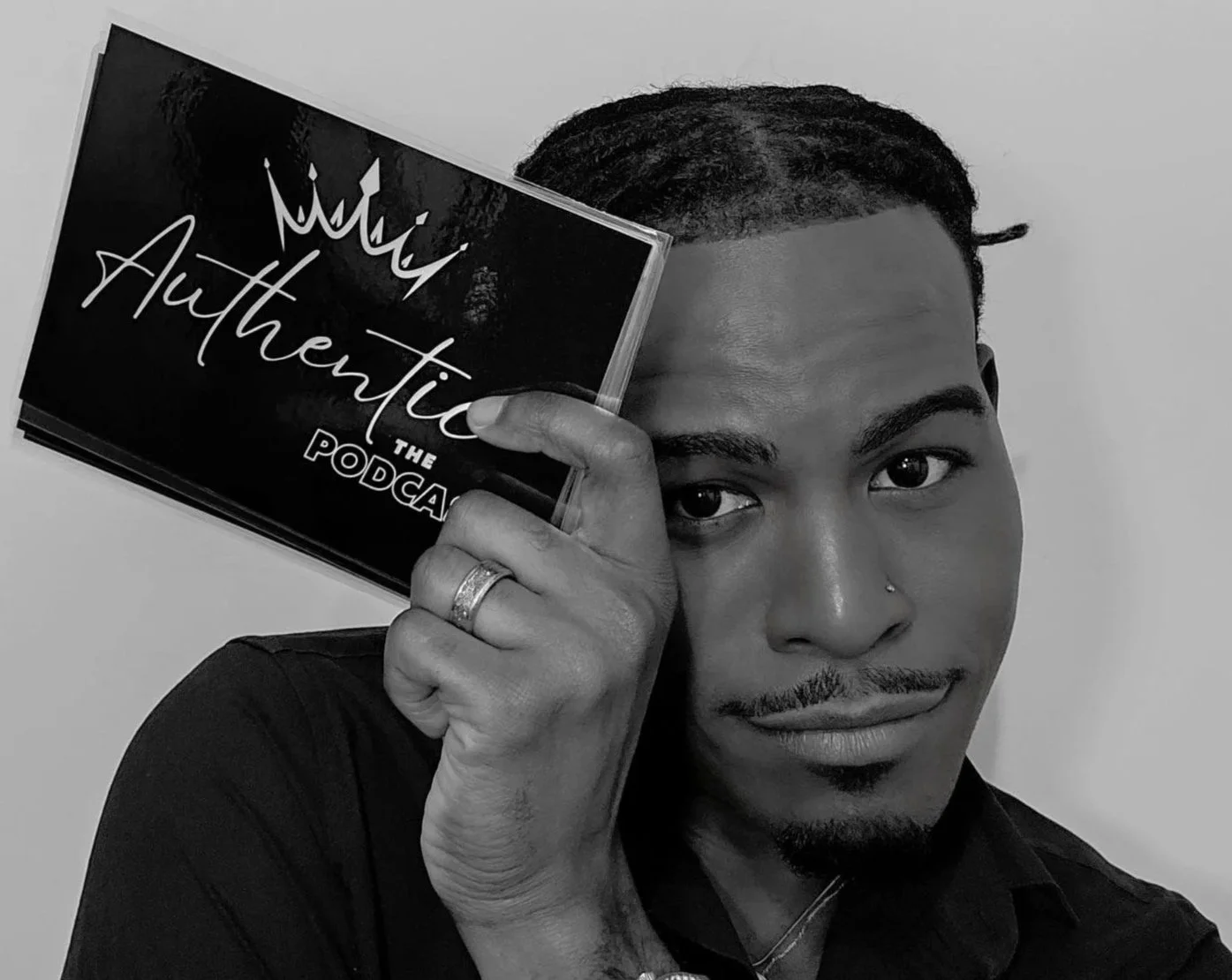 A man with short hair and a nose piercing holding a podcast cover titled "Authentic The Podcast" with a crown illustration, covering one of his eyes, smiling at the camera against a plain background.