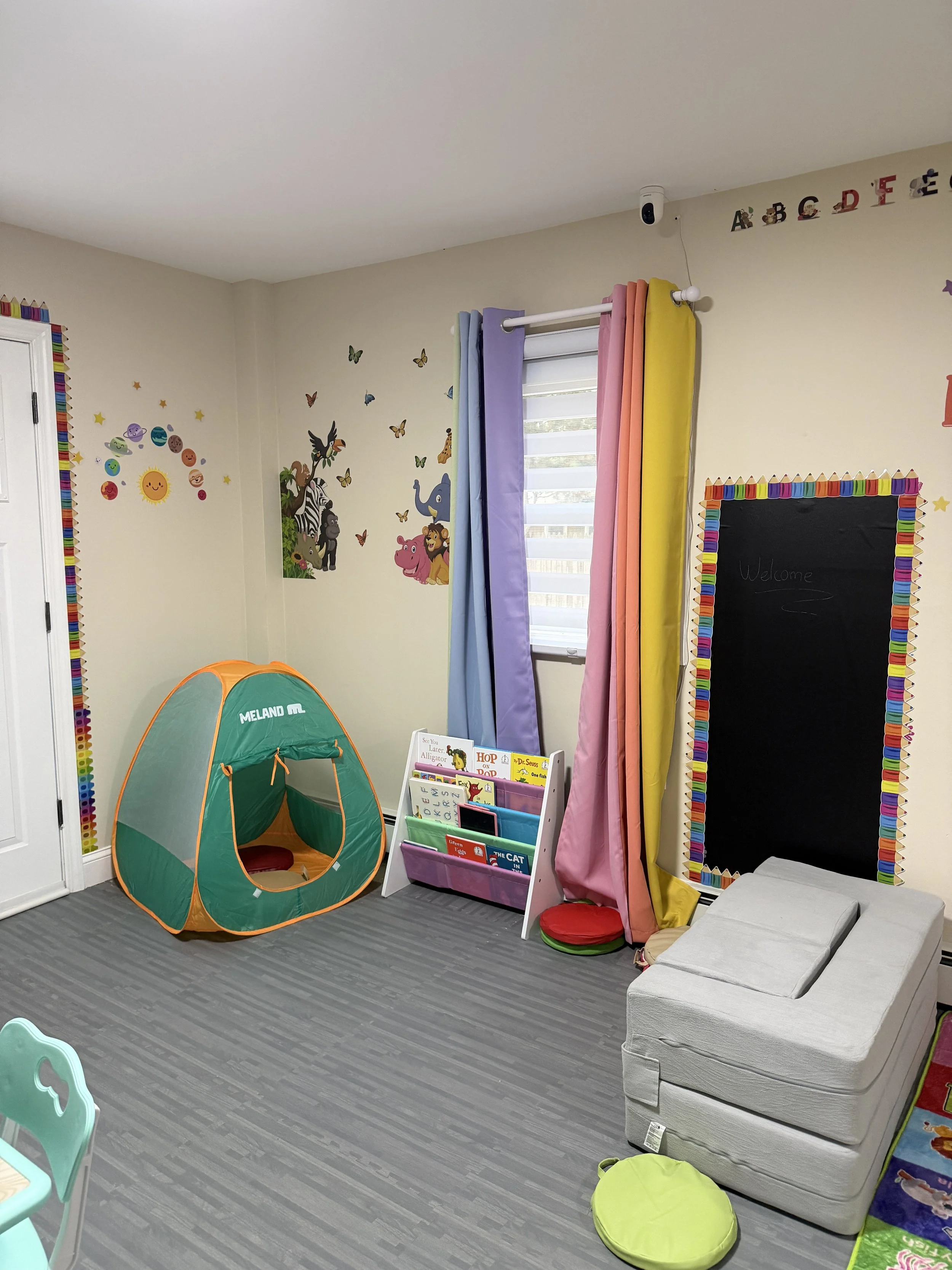 A colorful children's playroom with a small orange and green fabric play tent, a book display with children's books, a set of rainbow-colored curtains, a blackboard with a rainbow border, and a white couch against the wall with various wall stickers including animals and the alphabet.