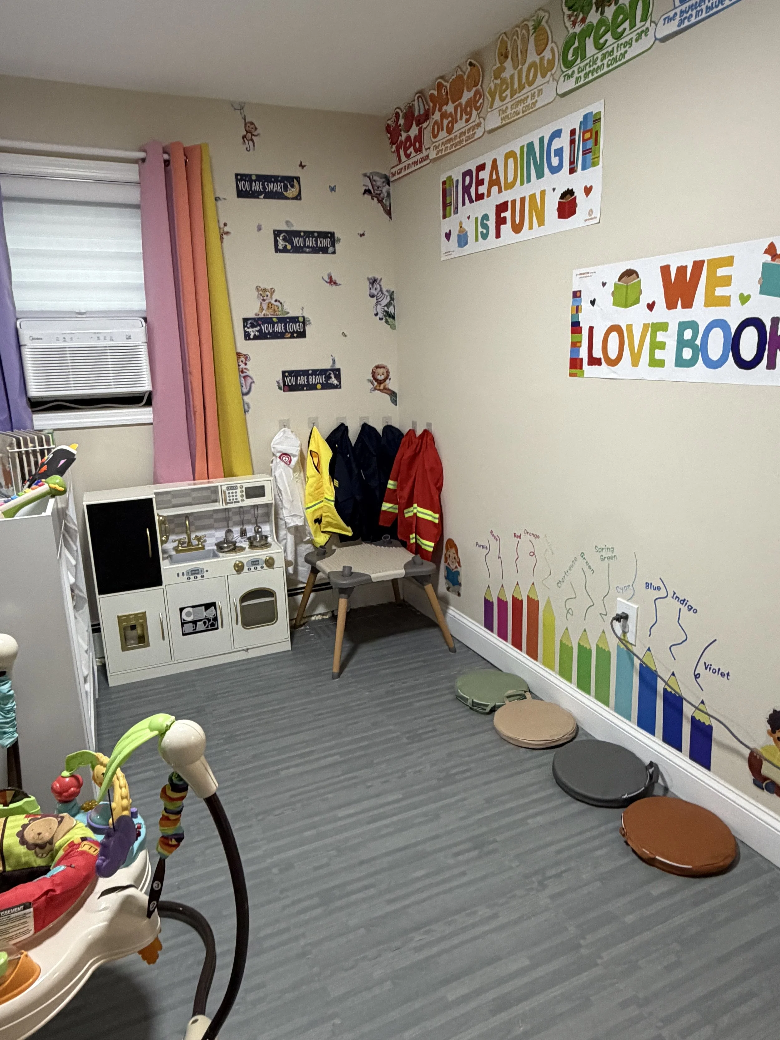 A corner of a children's room with educational posters, a small play kitchen, and colorful raincoat hangers, with two circular cushions on the floor and a baby walker in the foreground.
