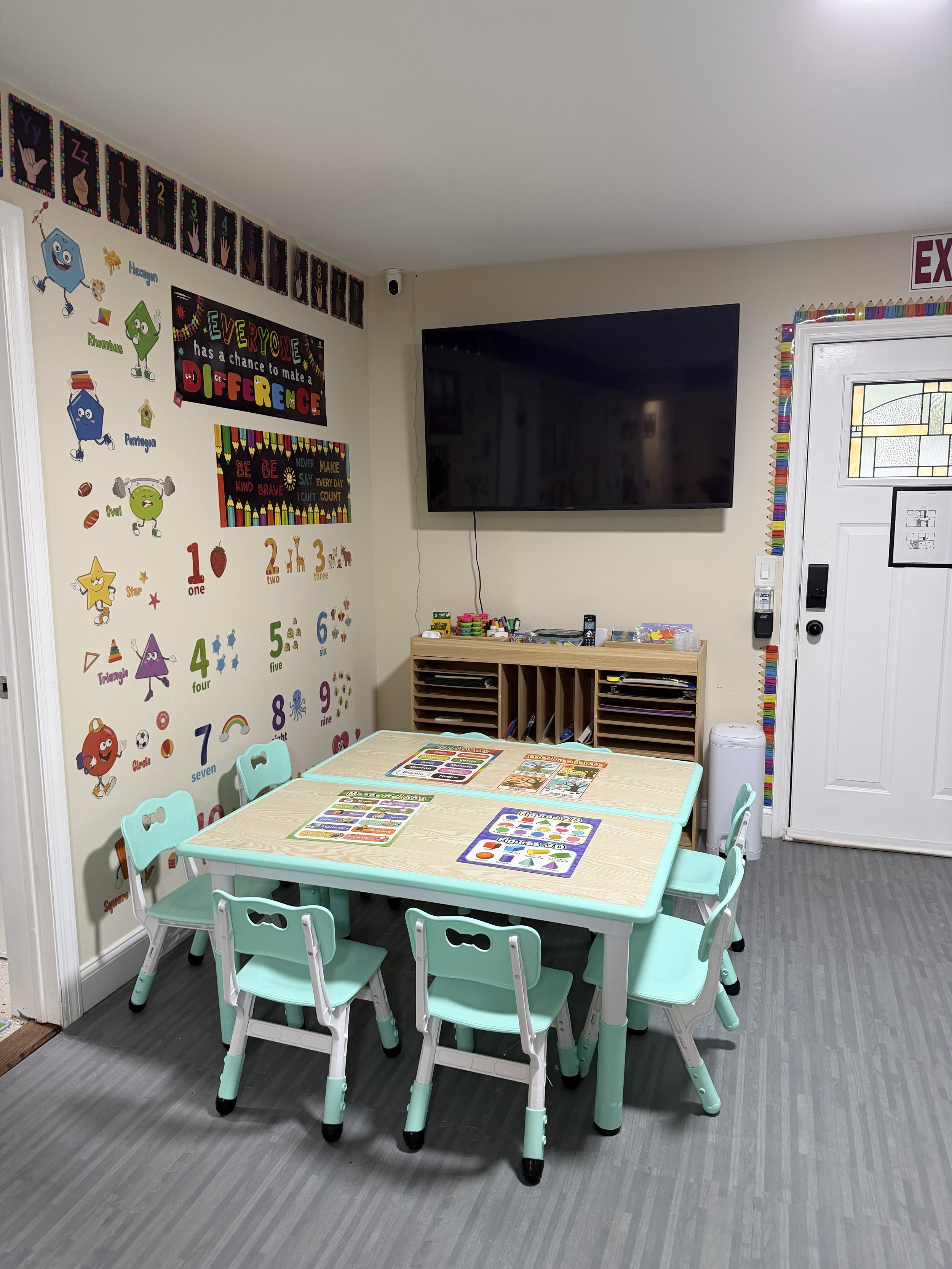 Classroom with educational posters on the wall, a black TV mounted on the wall, and a small light-colored table with turquoise chairs around it, with learning activity sheets on the table and colorful decorations around the door.
