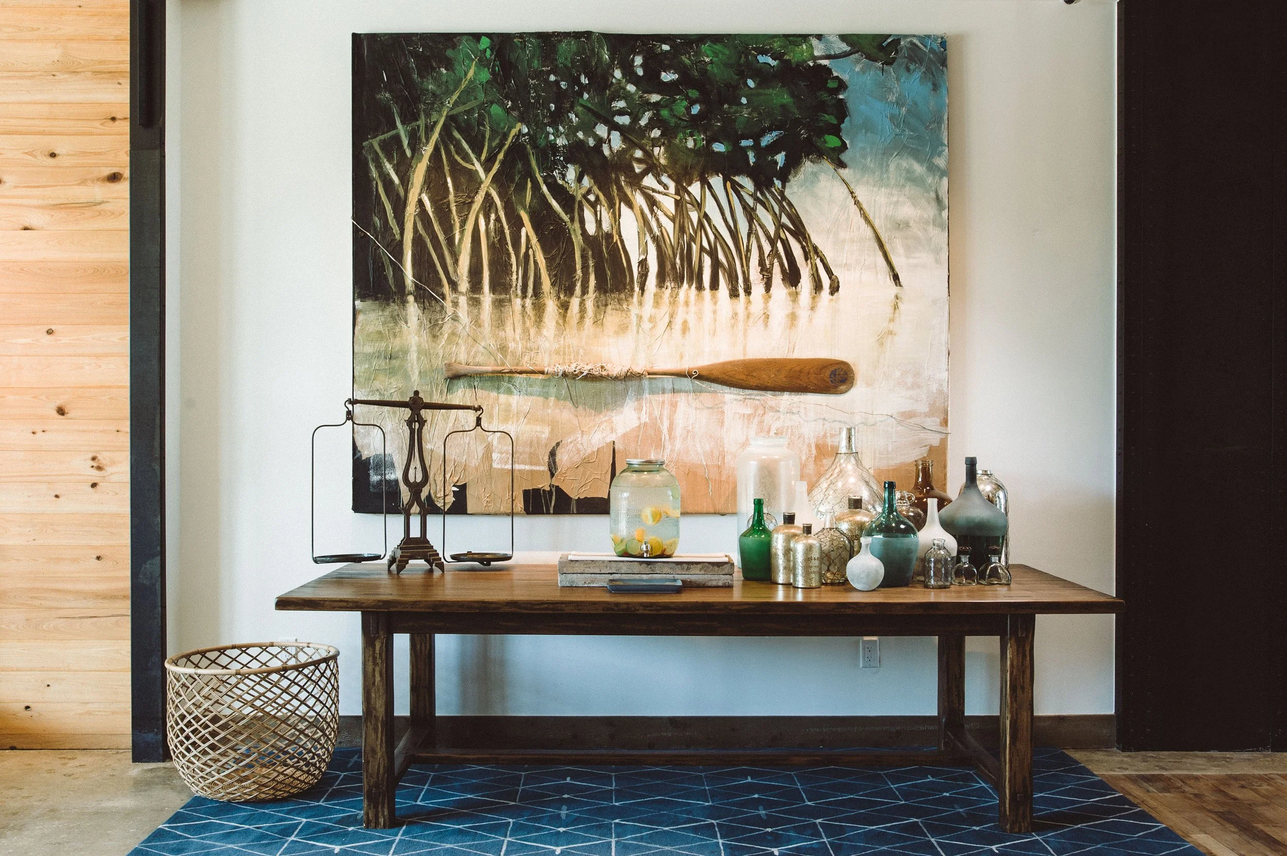A wooden table with various glass bottles and jars, a scale, and a painting of mangroves hanging on the wall behind the table. There is a blue geometric rug underneath the table and a woven basket on the floor to the left.