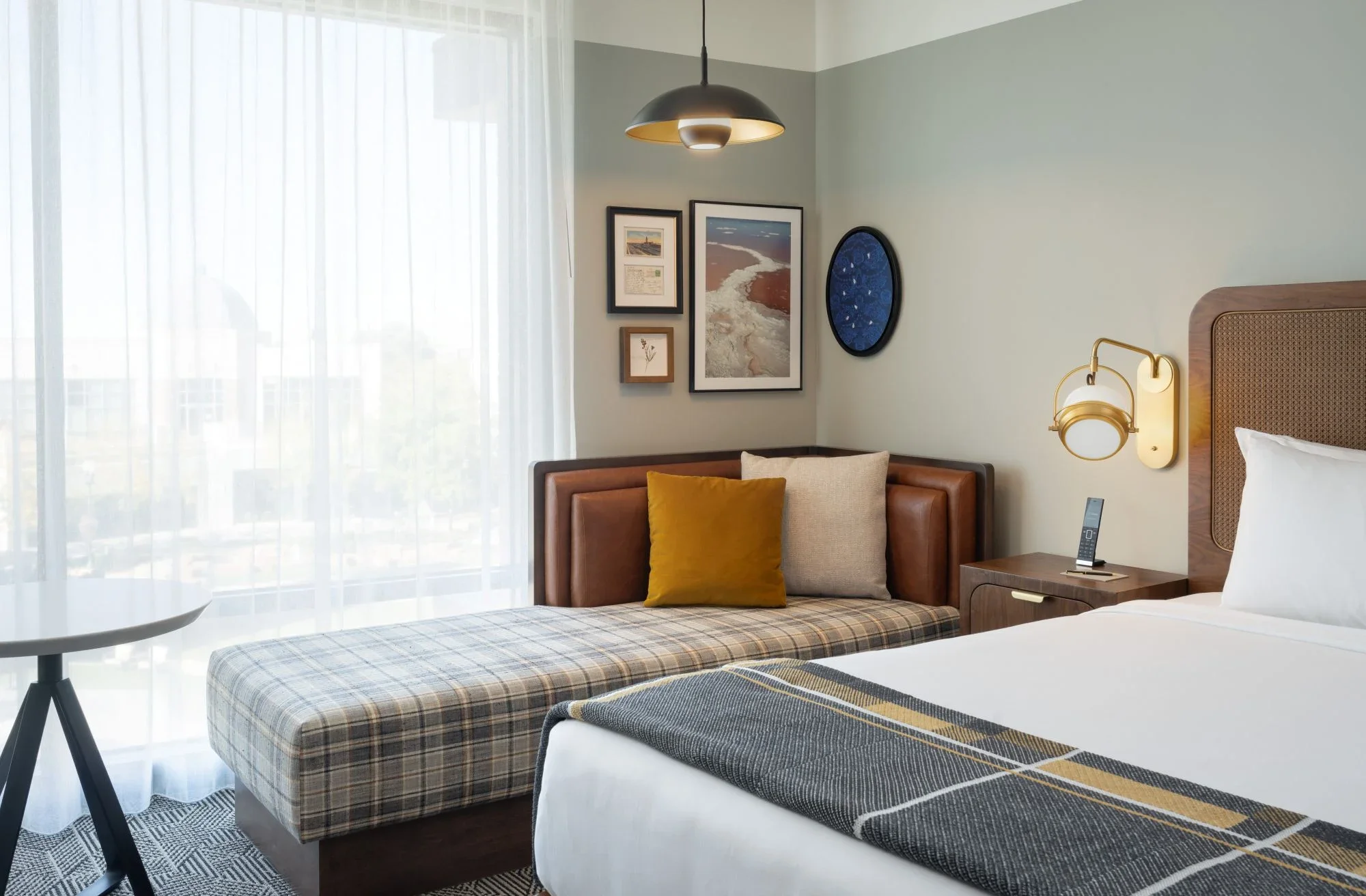 A hotel room with a bed, a plaid bench at the foot of the bed, a couch with a yellow throw pillow, a nightstand with a cordless phone, a wall-mounted light fixture, and framed artwork on the wall near a window with sheer curtains.