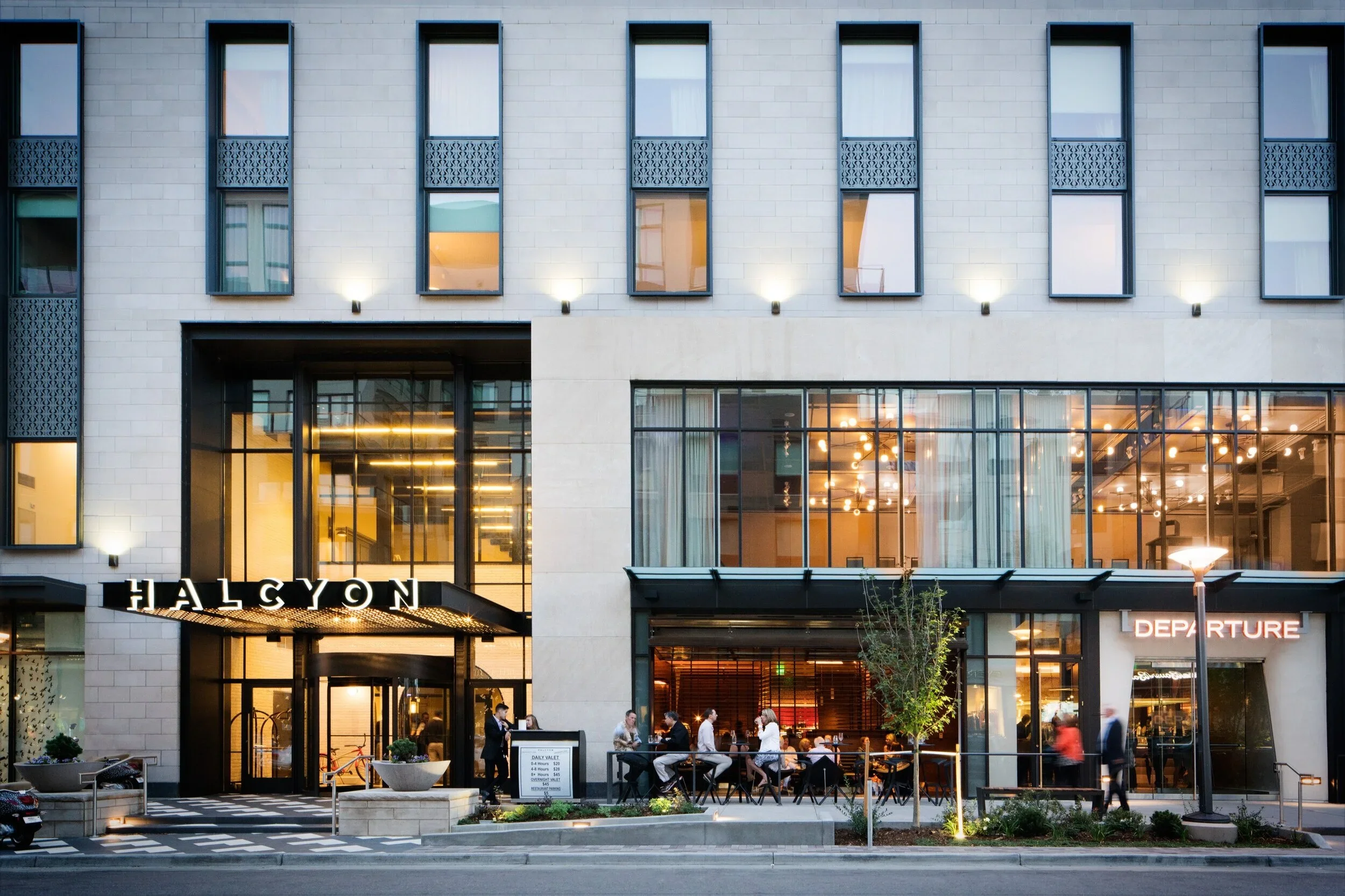 Modern building with large glass windows, people dining outside, illuminated sign reading "HALCYON," and a street scene at dusk.