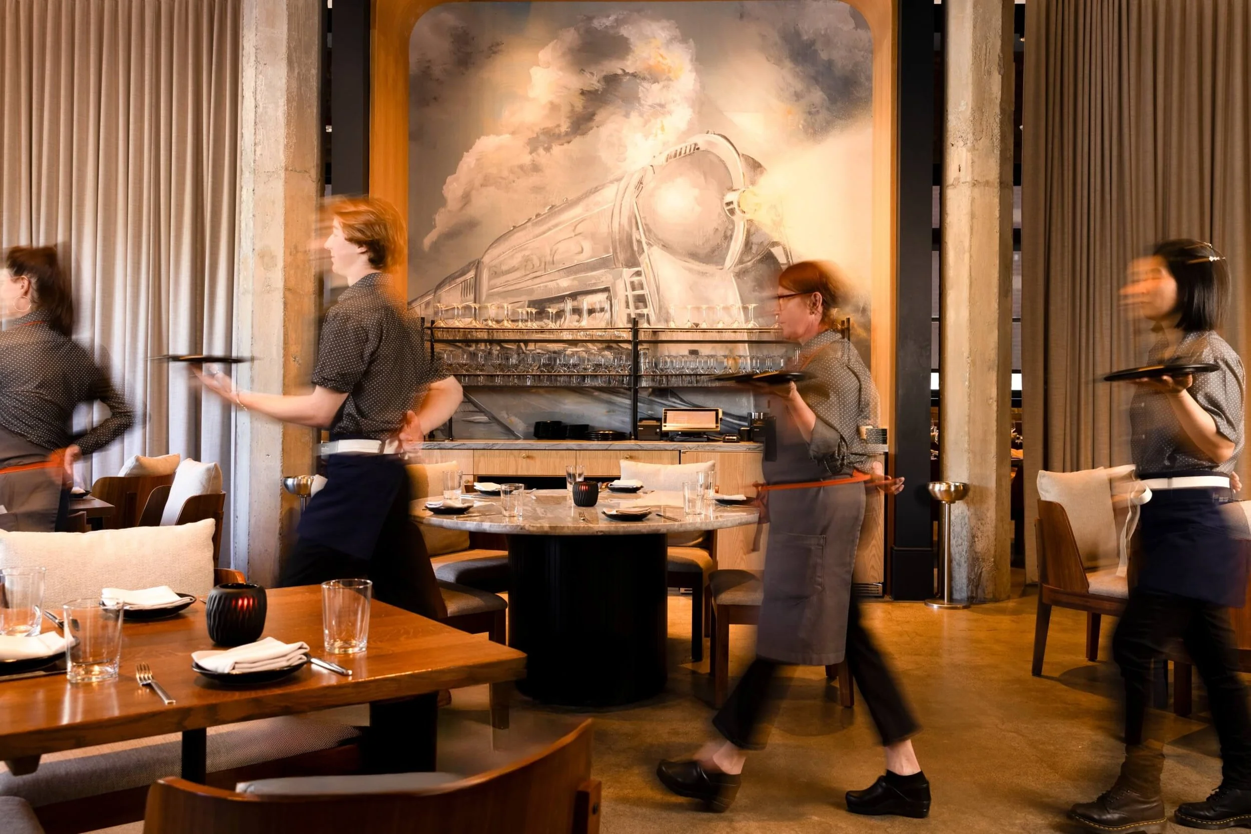 A restaurant interior with waitstaff carrying trays, a large artwork of a train on the wall, and dining tables set with glasses, plates, and utensils.