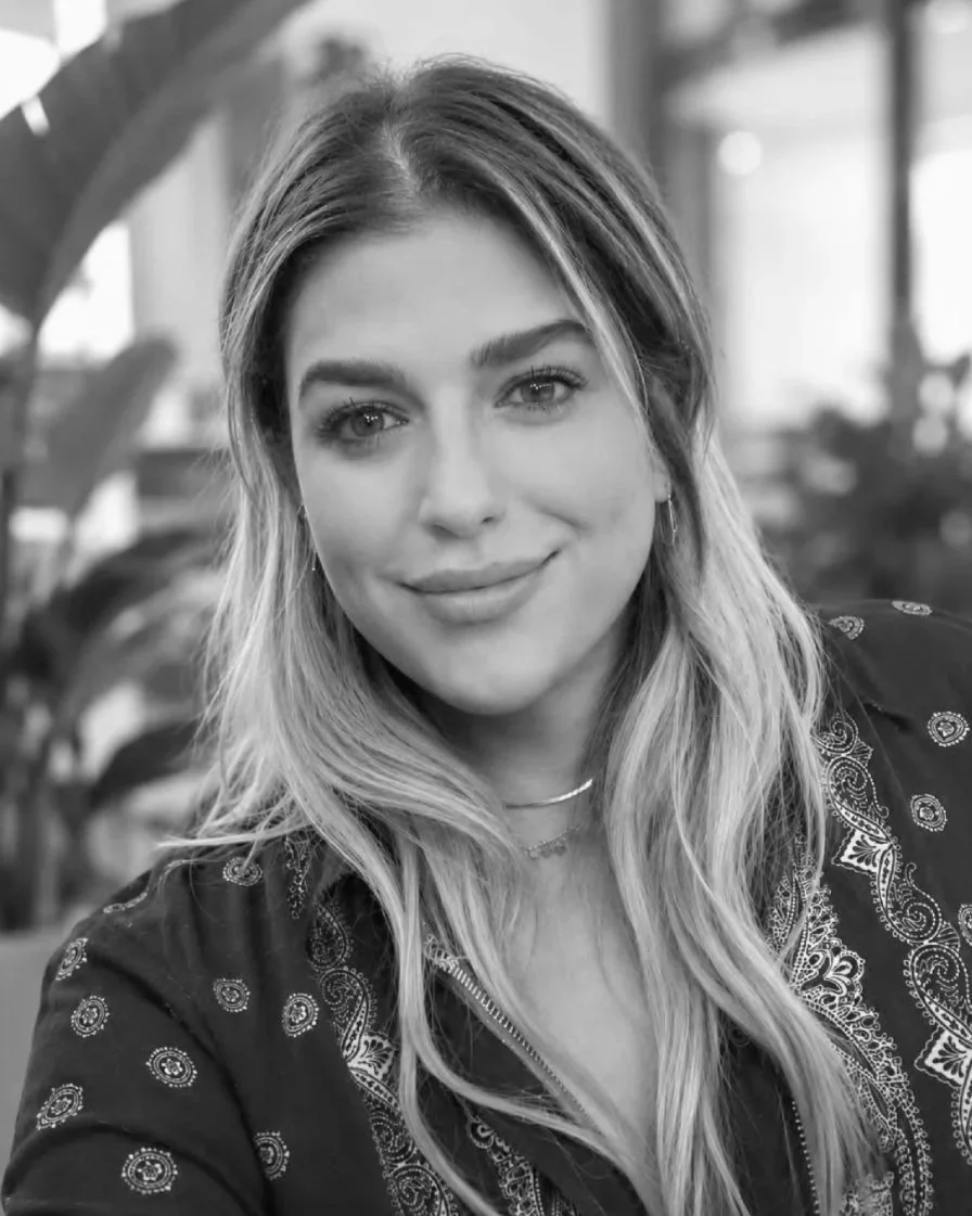 Close-up of a young woman with long, wavy hair and a gentle smile, wearing a patterned jacket and layered necklaces, in an indoor setting with plants and windows in the background.