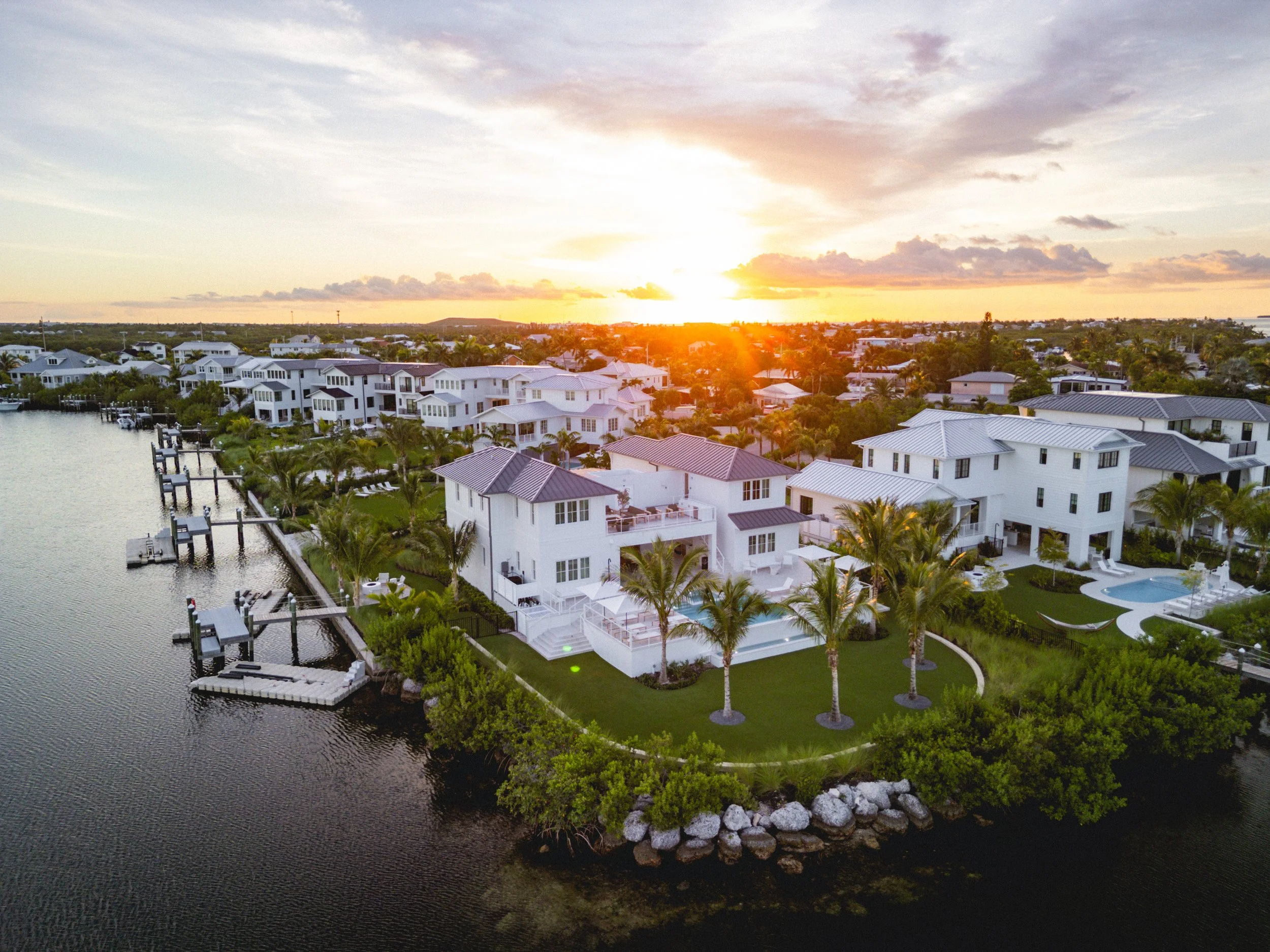 Aerial view of a waterfront residential neighborhood at sunset with several modern white houses, palm trees, a dock on a waterway, and a partly cloudy sky.