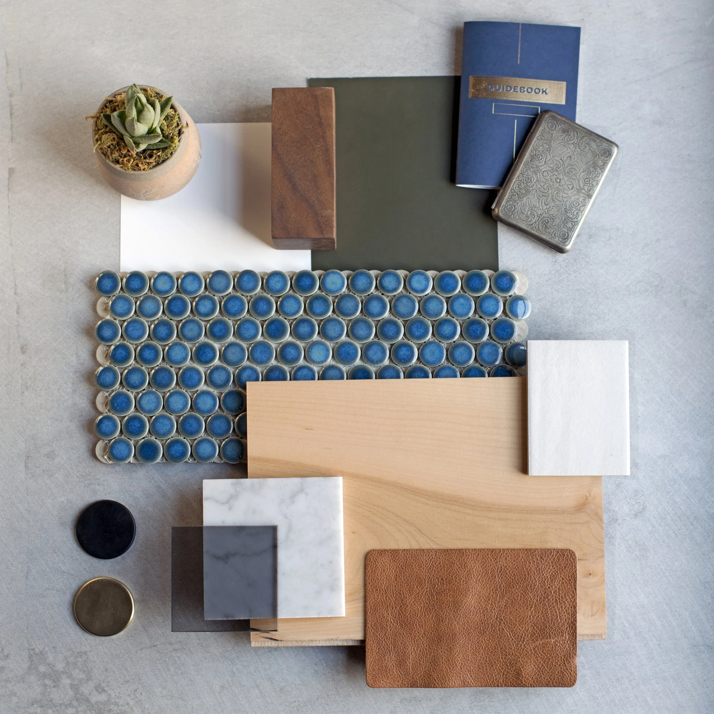 A flat lay of various objects including a small potted succulent plant, a brown rectangular block, more than three books including a blue guidebook, a dark green book, and a white book, a decorative metal box, a string of blue buttons, a black circul
