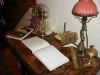 A wooden table with a white open notebook, a pink table lamp, a glass bell jar, and decorative items.