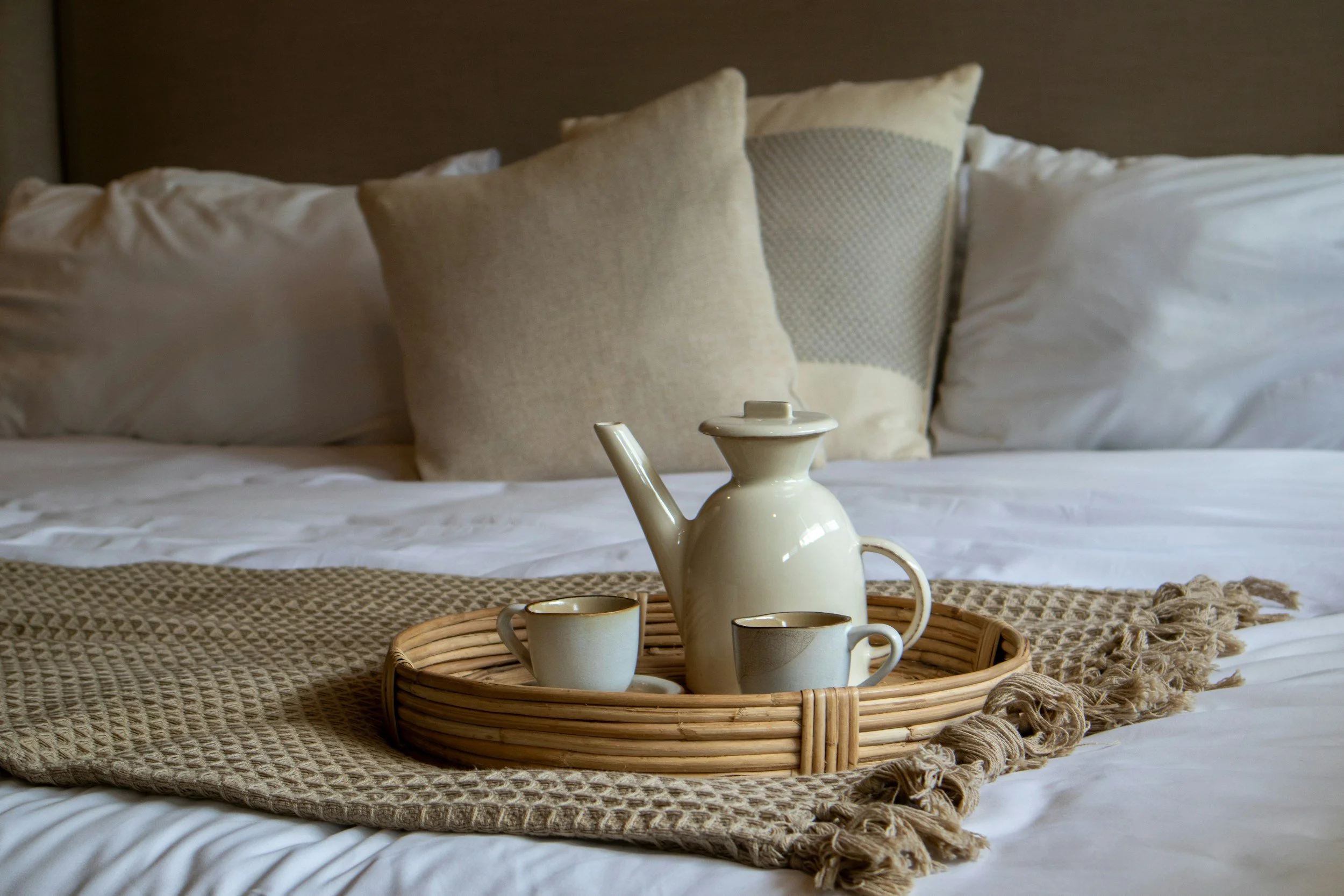 Postpartum doula care/support in San Antonio.  A cozy bed with white sheets, beige pillows, and a dark headboard, topped with a round wicker tray holding a cream-colored teapot and two matching cups, with a woven beige blanket at the foot of the bed.