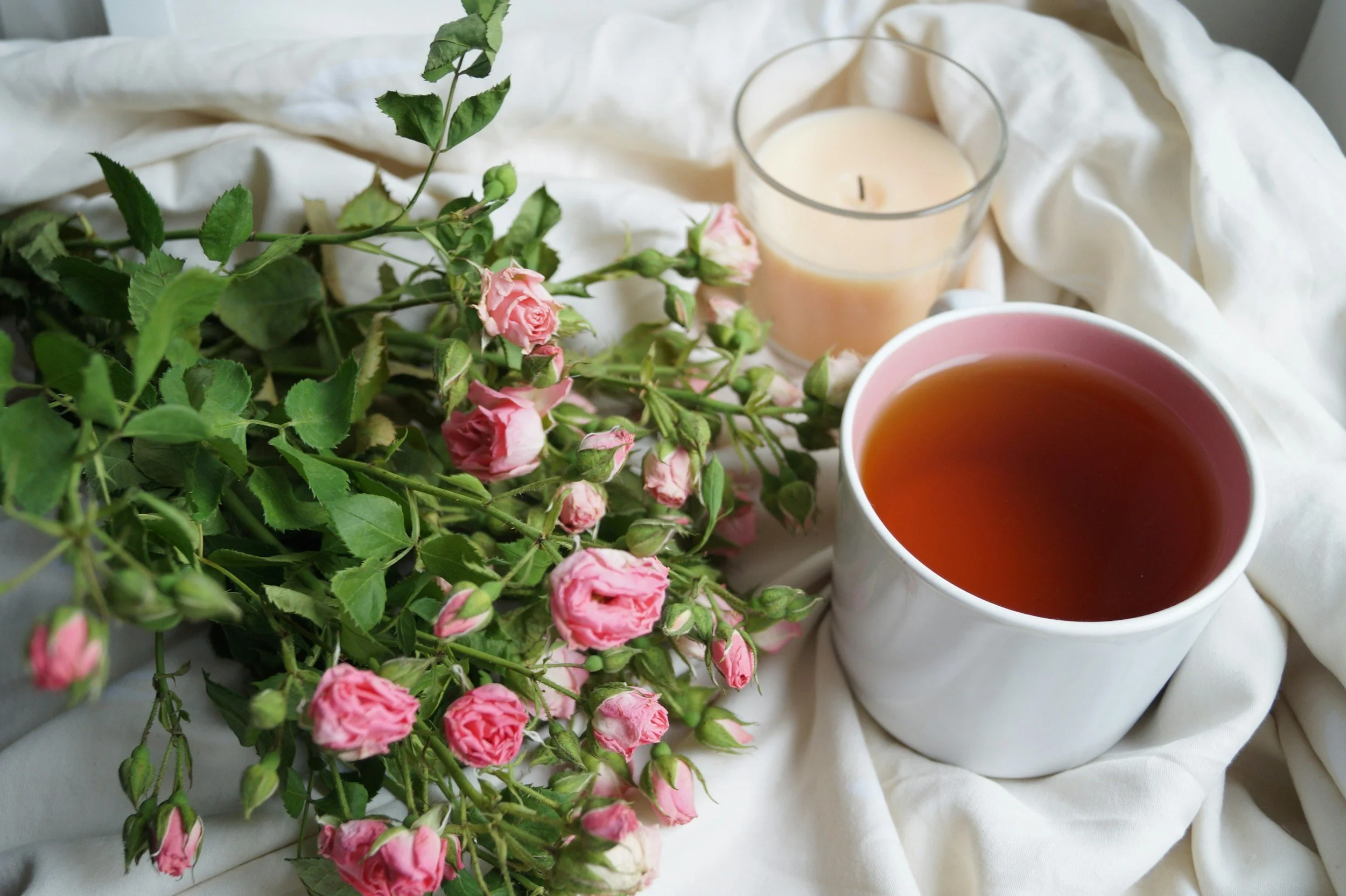 Bereavement photo: Compassionate pregnancy loss and bereavement doula support. Pink roses with green leaves, a candle in a glass holder, and a cup of tea on a white fabric surface.