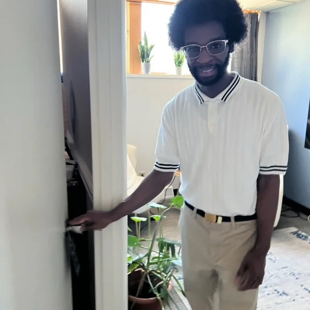 Picture of Andre he is a man with curly hair and glasses standing in a living room, holding a door handle, wearing a white collared shirt with black stripes, khaki pants, and a black belt.