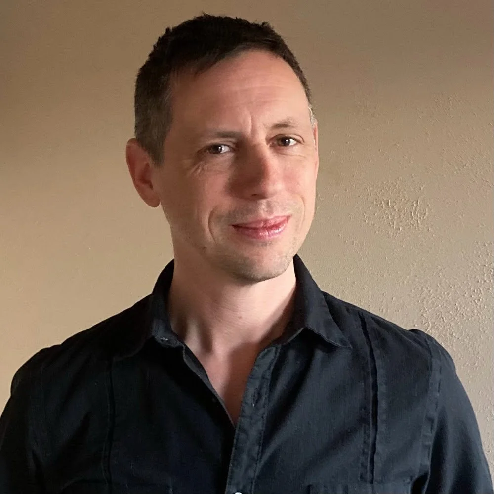 A man with short brown hair and light skin wearing a black collared shirt standing against a beige wall.