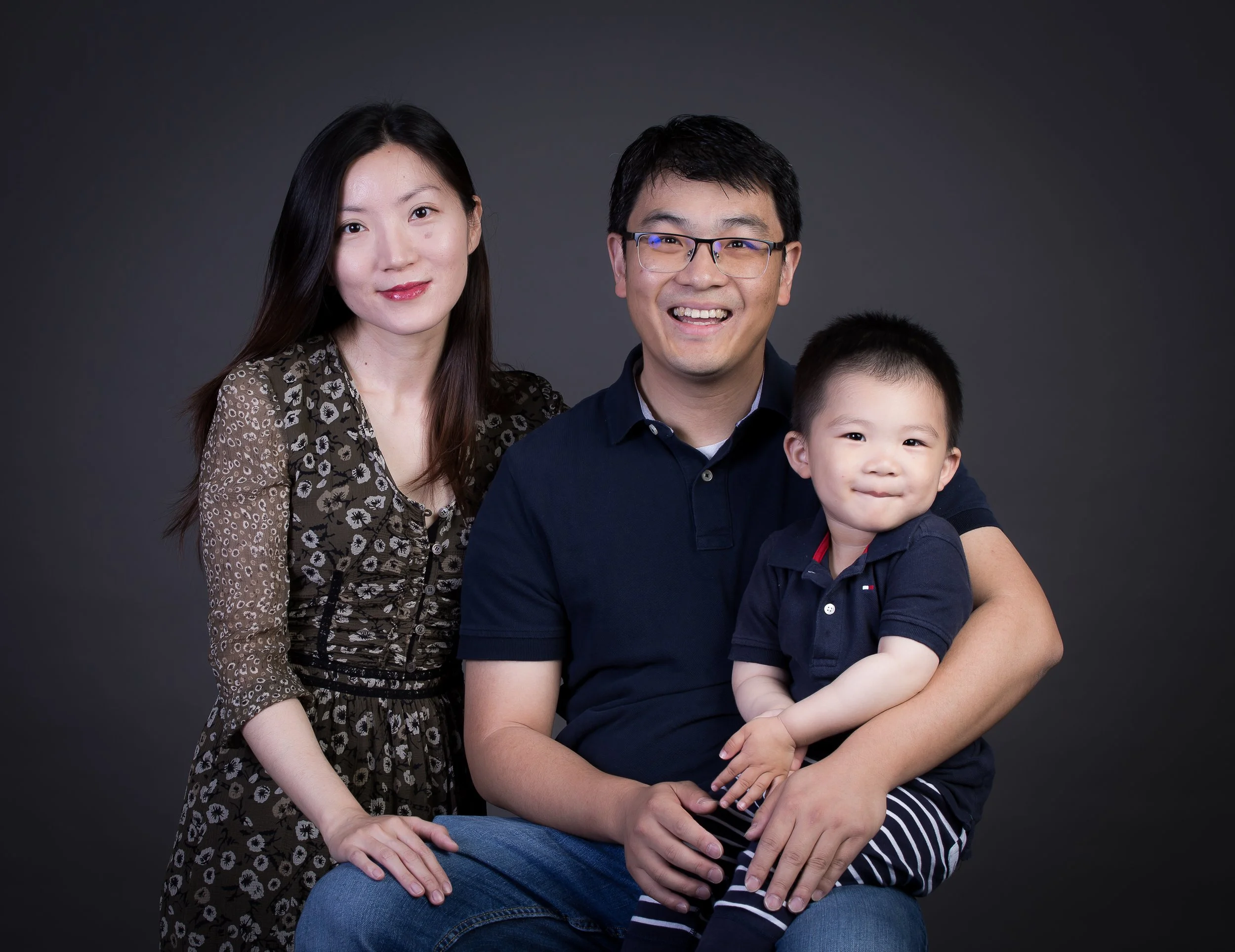 An Asian family of three posing for a portrait. The mother has long black hair and is wearing a patterned dress, the father has short black hair with glasses and is smiling, holding their young son who has short black hair and is wearing a navy blue 