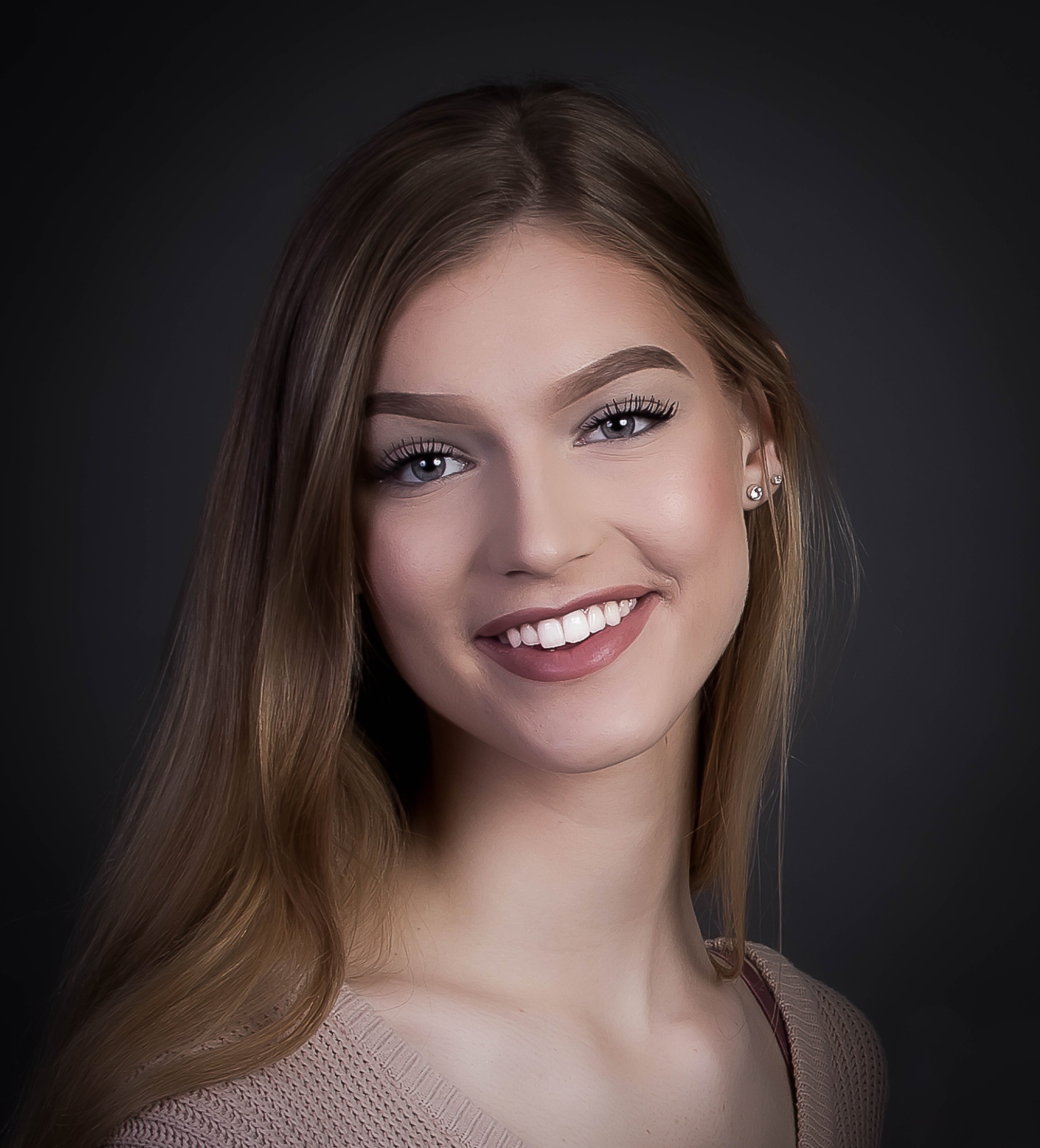 Close-up portrait of a young woman with long, brown hair, smiling, against a dark background.