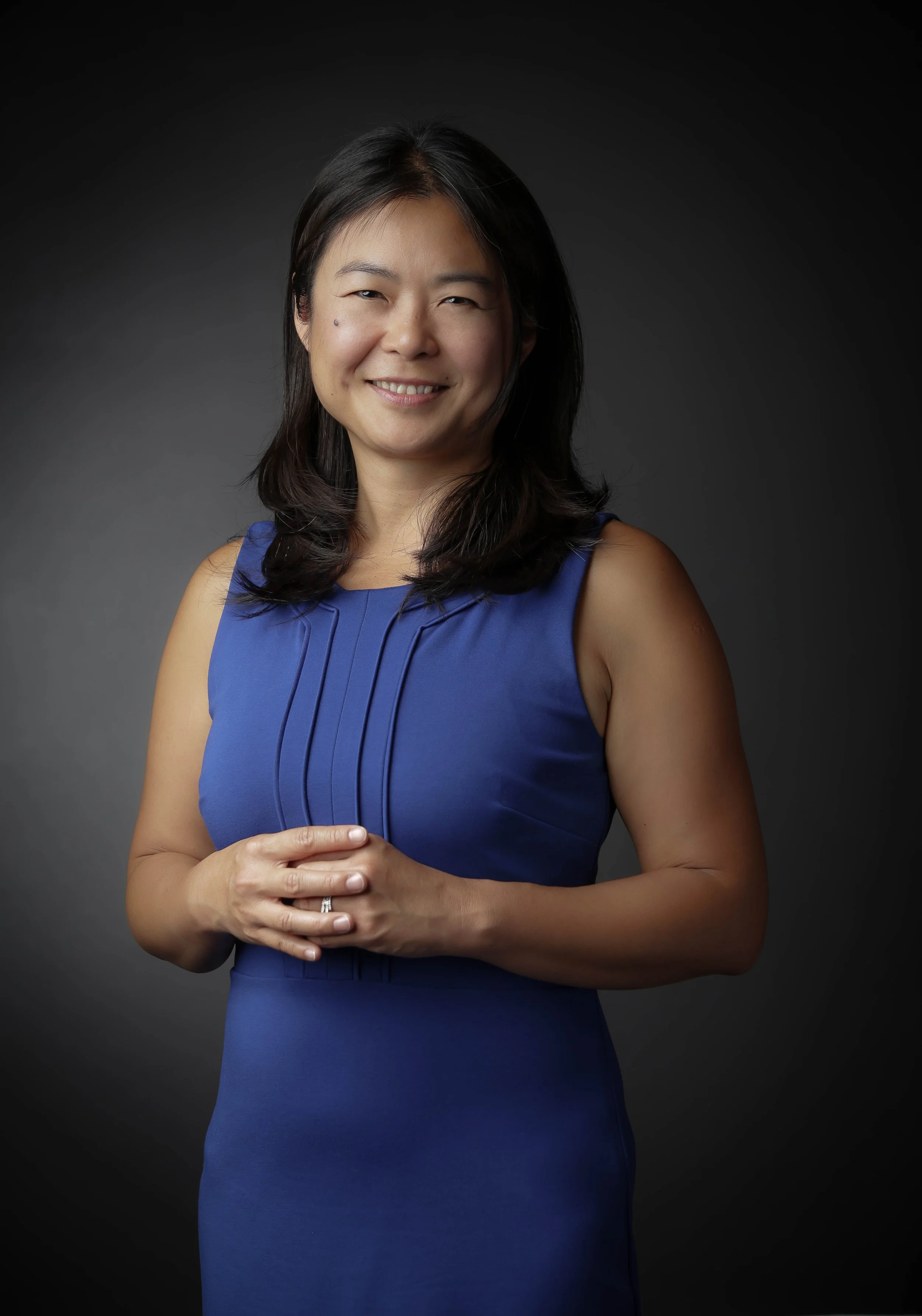 Portrait of an Asian woman wearing a sleeveless blue dress, smiling, with her hands clasped at waist level, standing against a dark background.