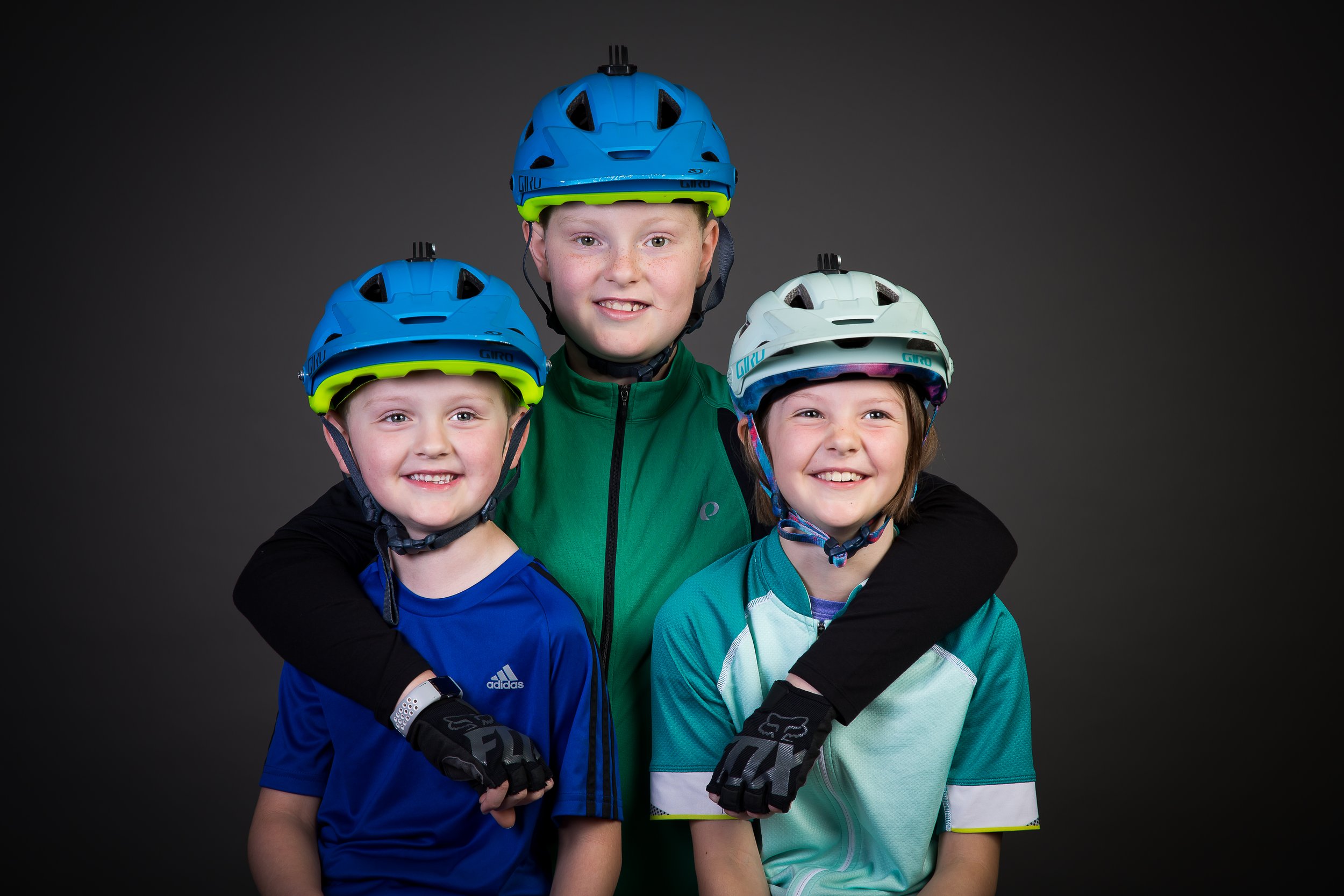 Three children wearing biking helmets and gloves, smiling at the camera, against a dark background.
