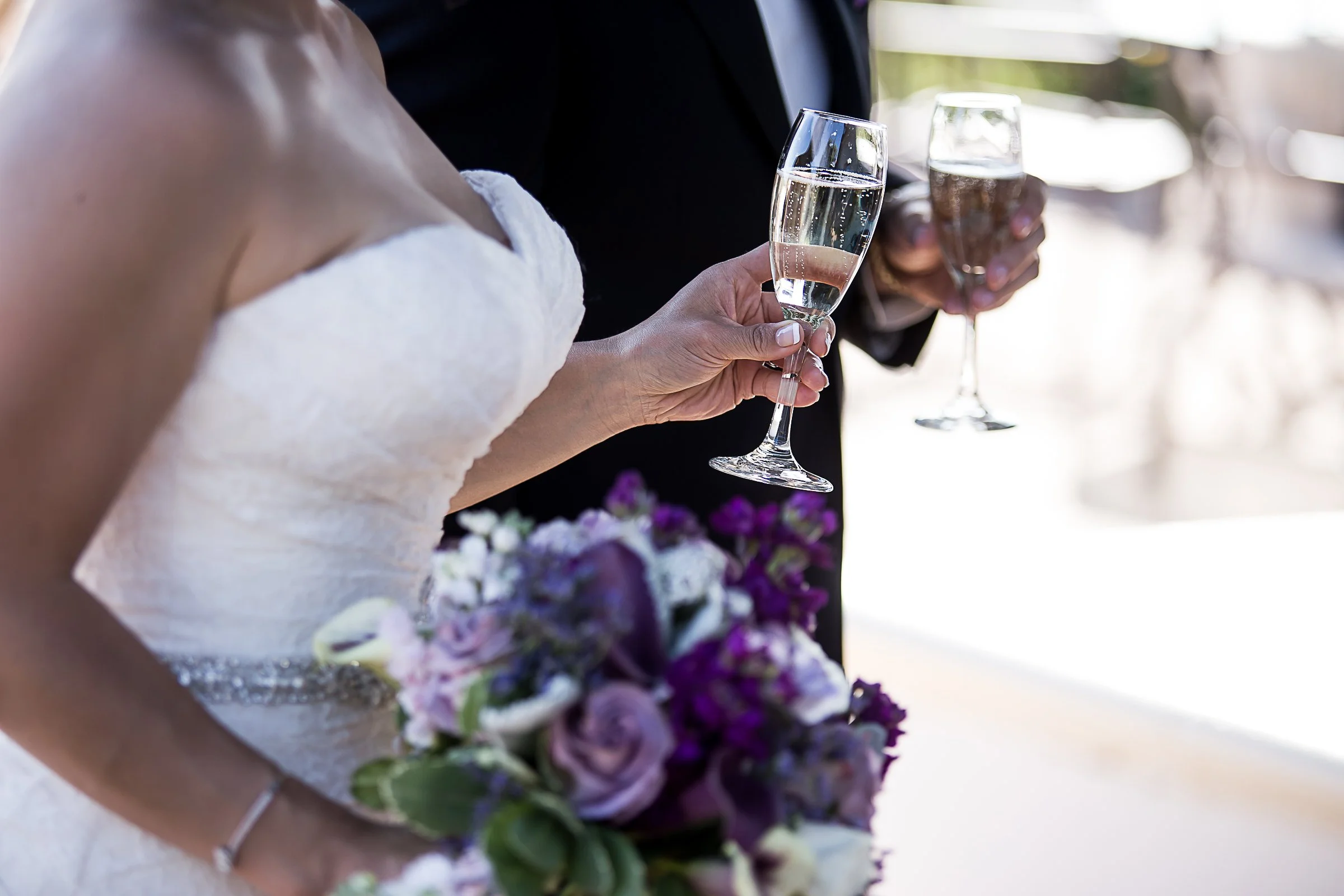 A bride and groom at a wedding, holding glasses of champagne over a table decorated with purple and white flowers.
