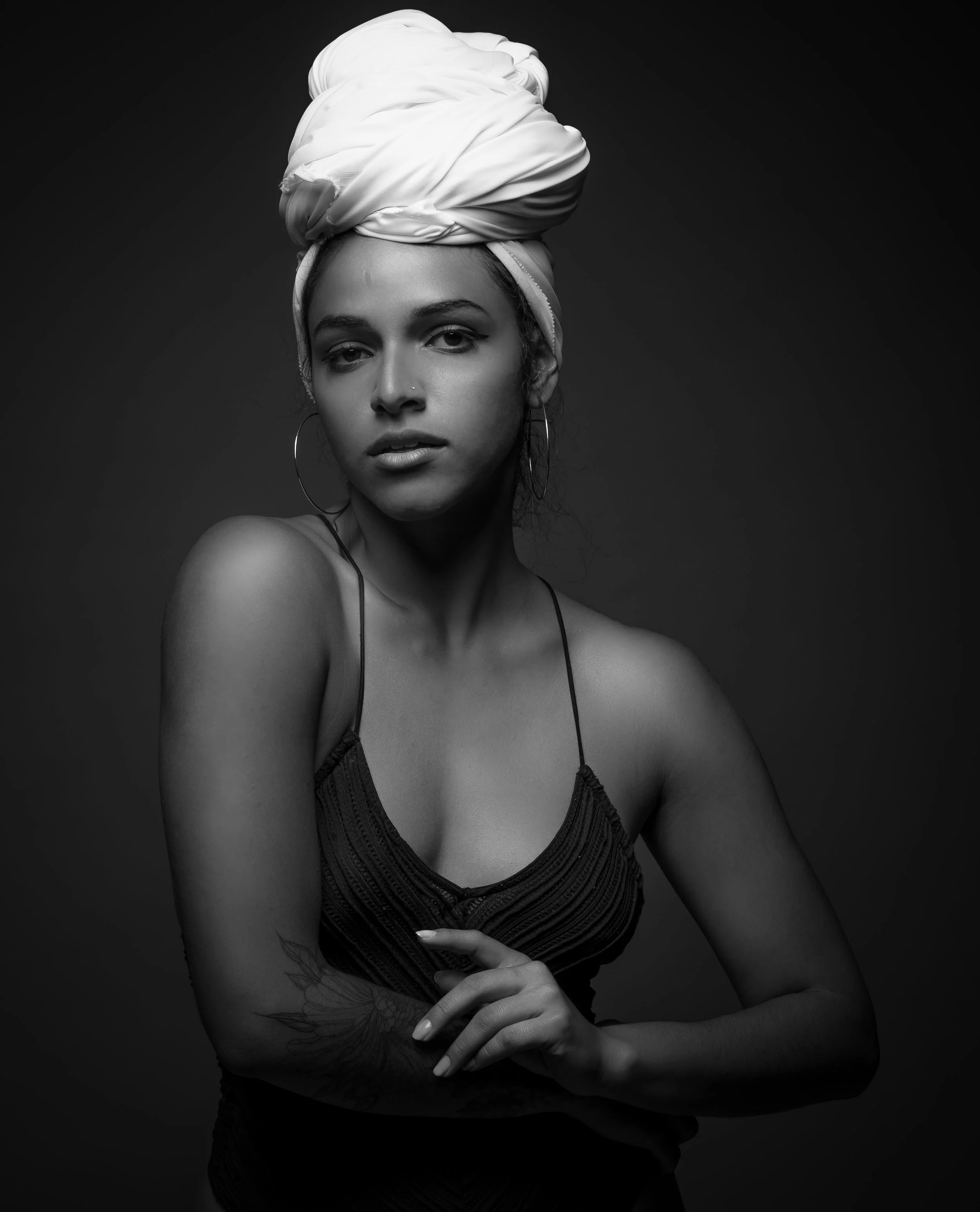 A woman with a dark headscarf wrapped in a large turban hold her hands together in front of her with a floral tattoo on her arm, wearing hoop earrings, against a dark background.