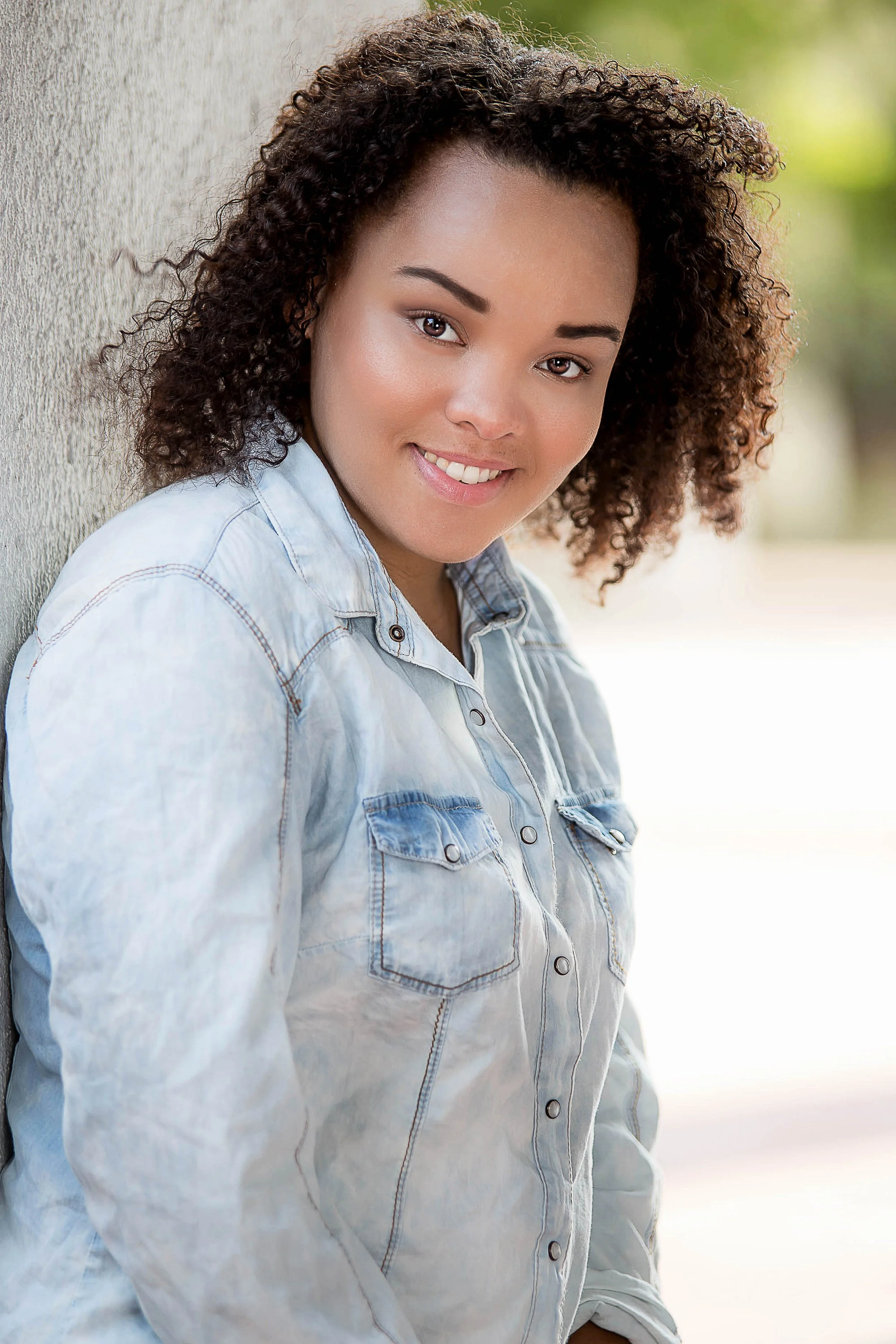 A young woman with curly hair and a light denim shirt smiling outdoors by a textured wall with a blurred green background.