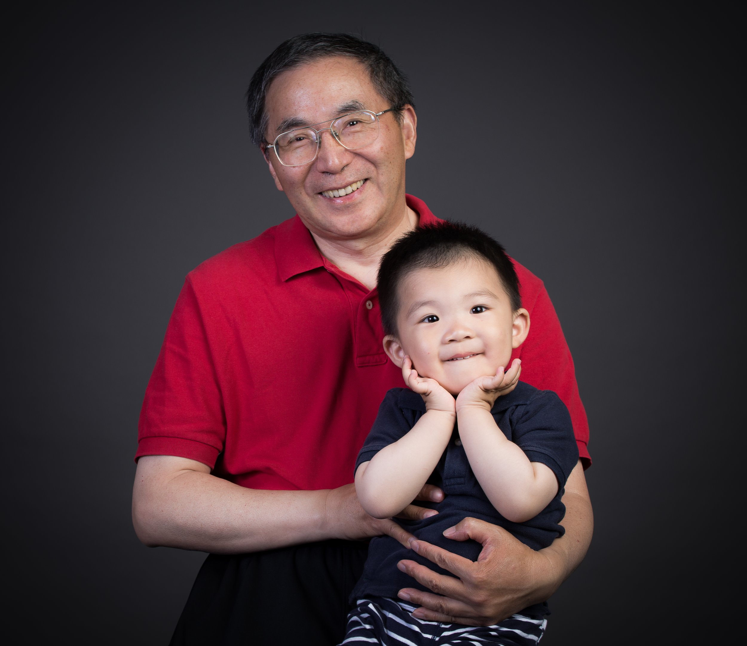 An Asian man in glasses and a red shirt smiling while holding a young boy in a navy shirt, against a dark grey background.