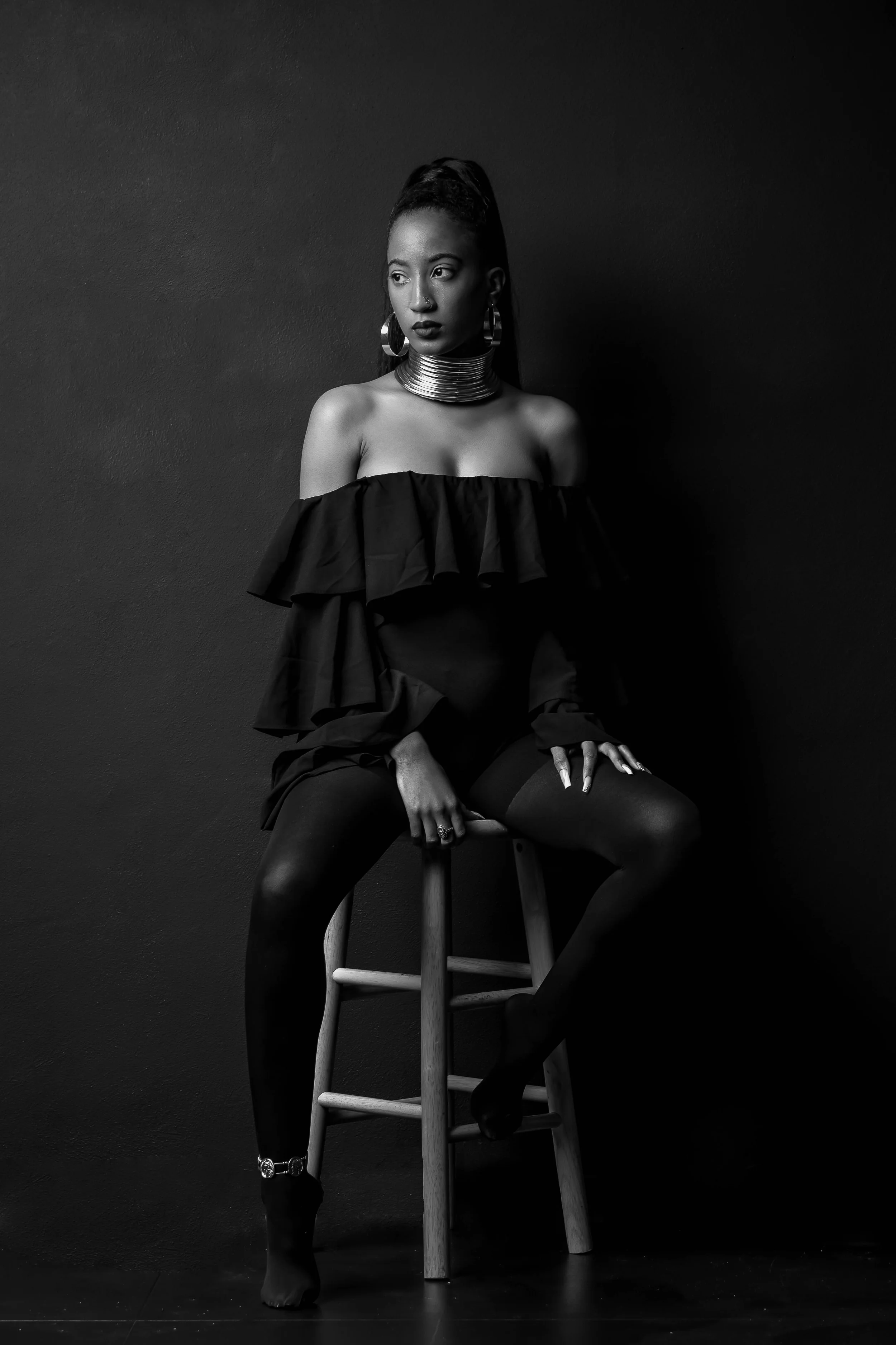 Black and white photo of a woman sitting on a wooden stool against a plain dark wall. She is wearing a ruffled off-the-shoulder top, black tights, and high heels with an ankle strap. She has jewelry including large hoop earrings, a nose ring, and mul
