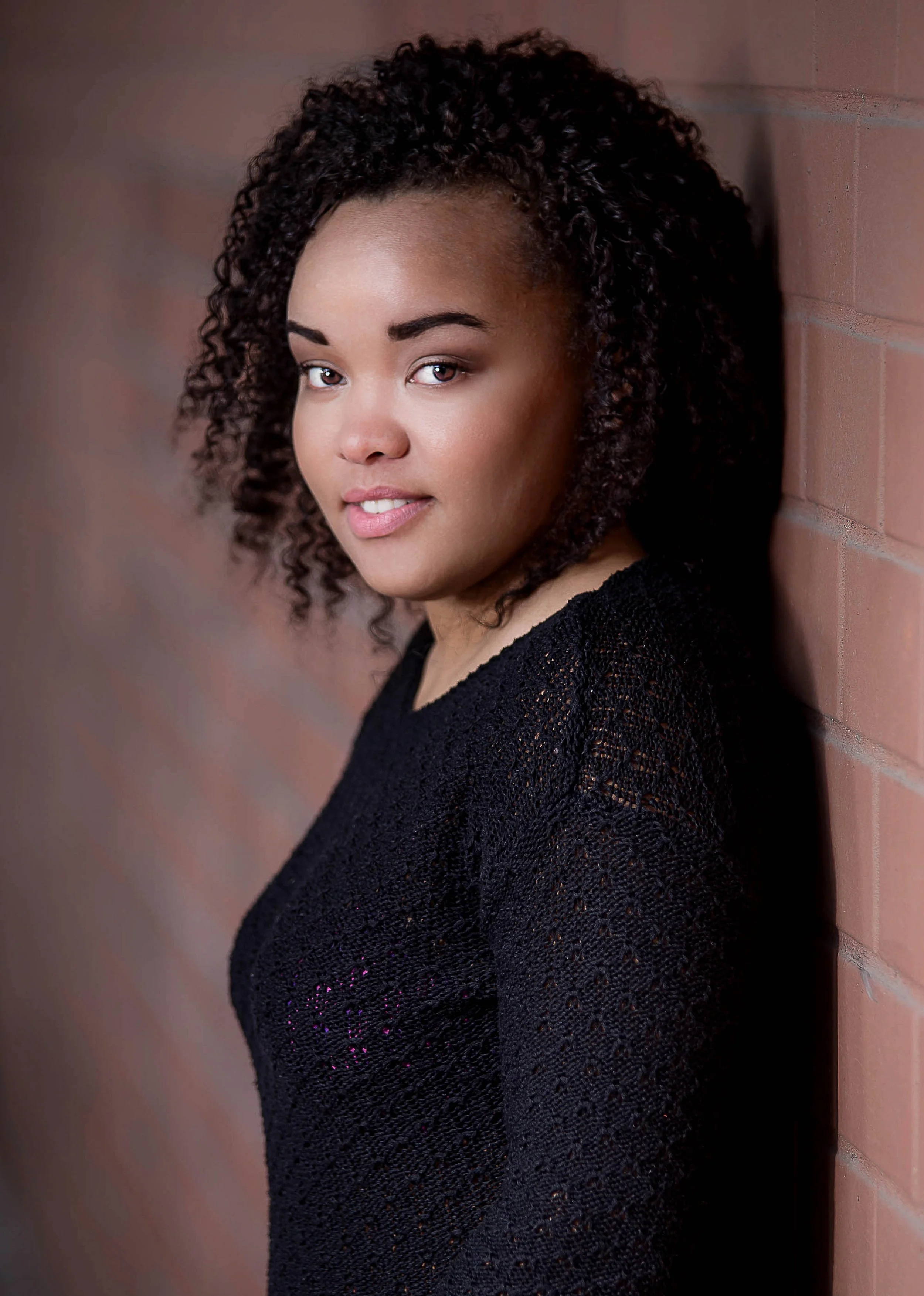 A young woman with curly black hair and a black top leaning against a brick wall, looking at the camera with a slight smile.