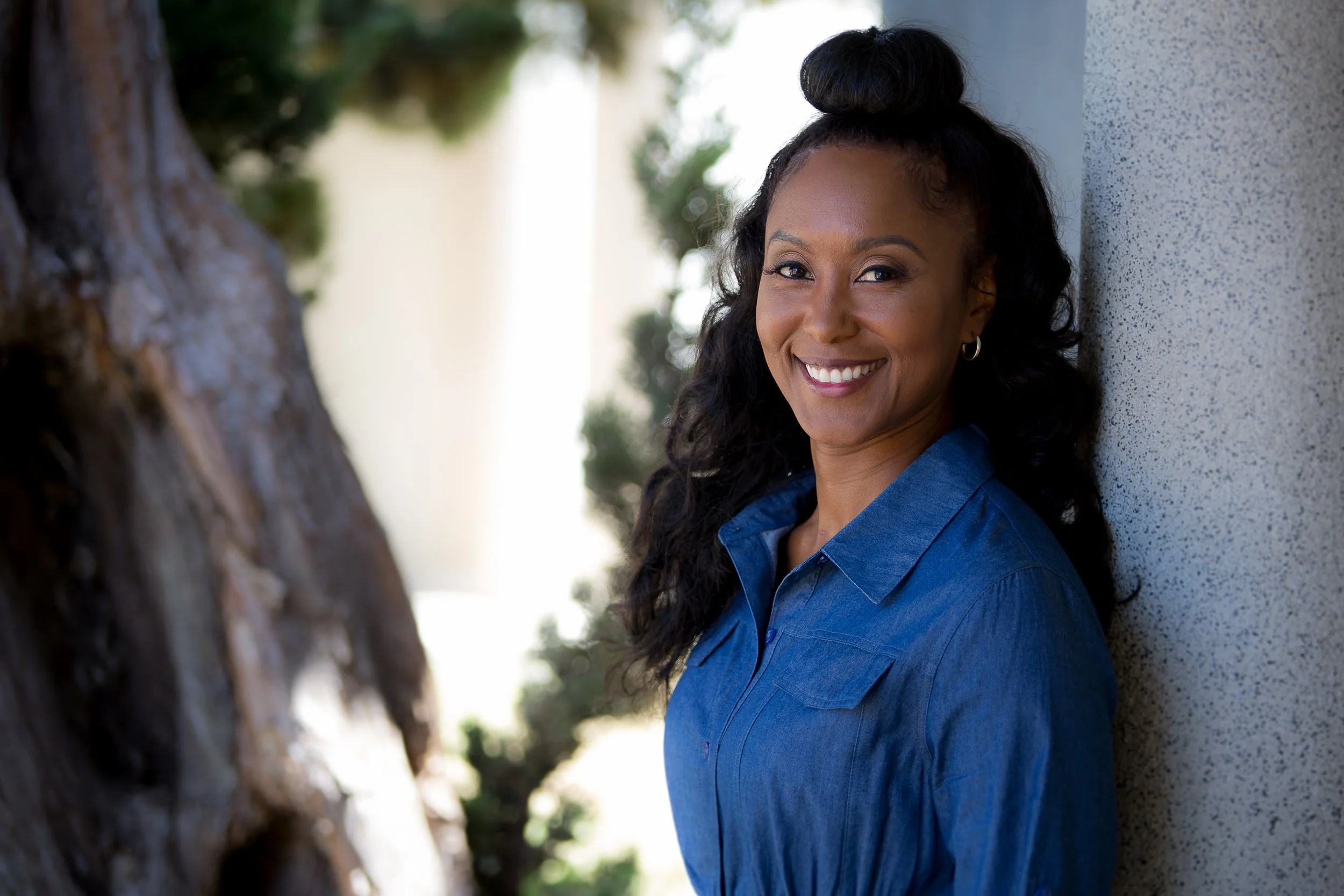 A woman with long curly hair styled in a top bun, wearing a blue denim shirt, smiling and leaning against a textured beige wall outdoors with trees blurred in the background.