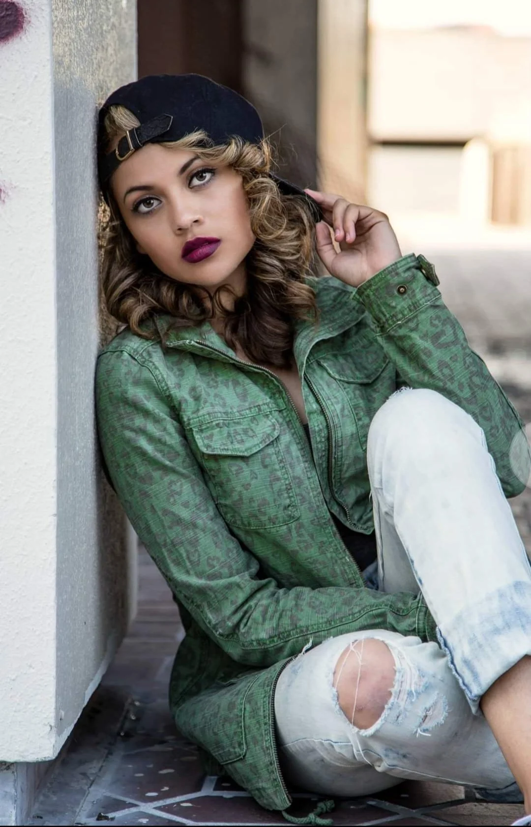 Young woman with curly hair wearing a black baseball cap backwards, green jacket, and ripped jeans, sitting on the ground against a wall, looking up with her hand near her face.