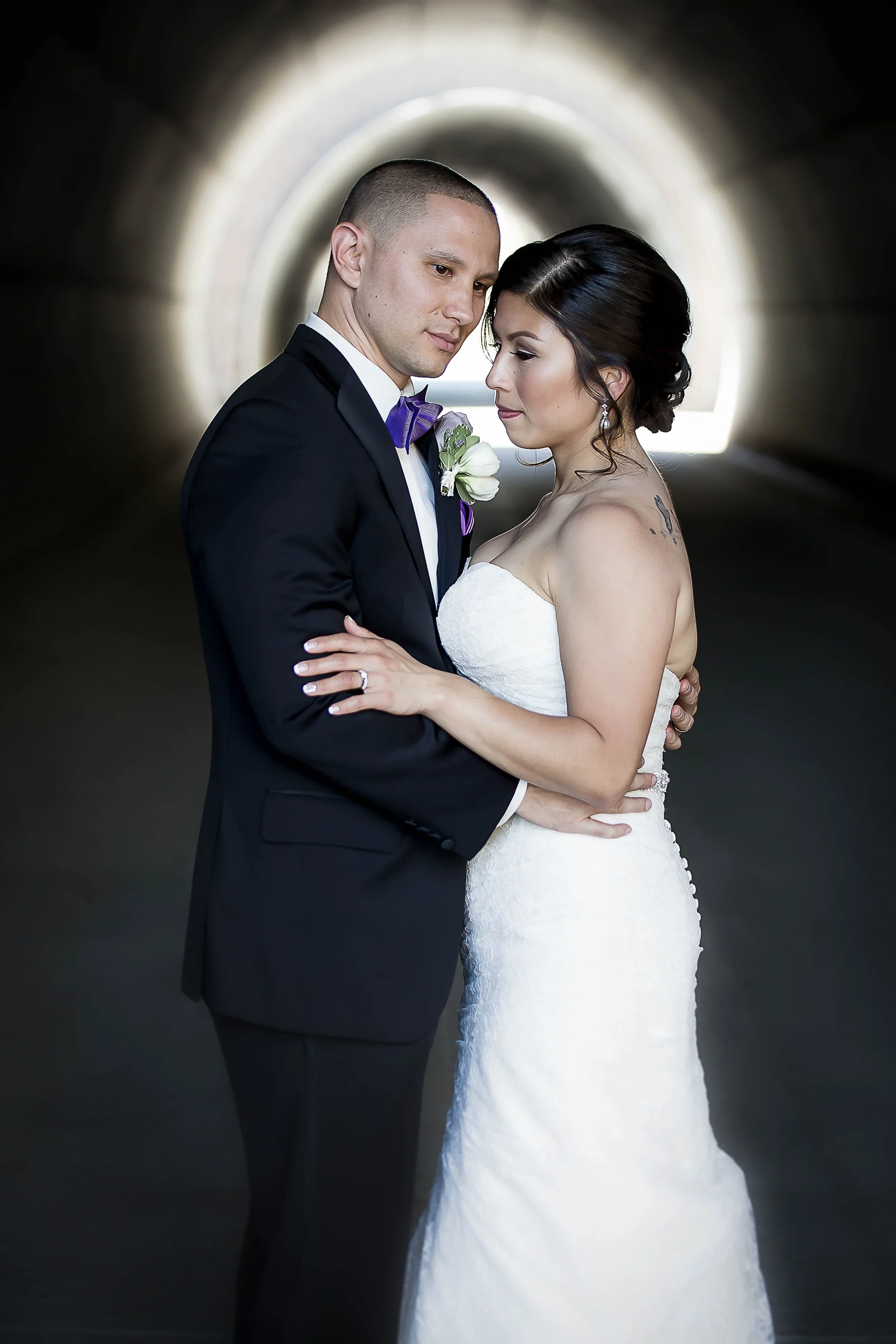 A bride and groom standing close together in a tunnel, with the bride wearing a white strapless wedding gown and the groom in a black tuxedo with a purple bow tie and boutonniere.