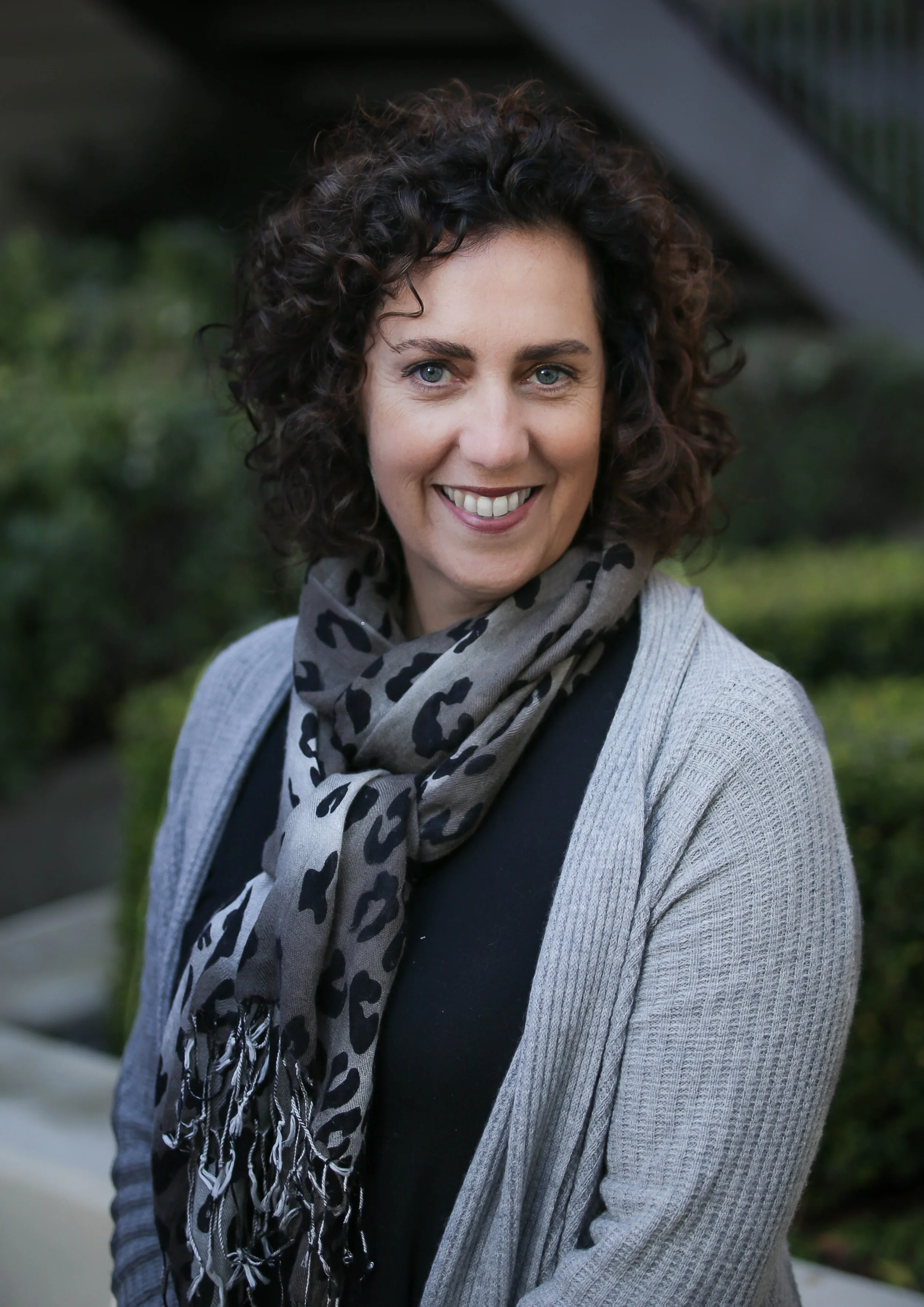 Woman with curly brown hair, wearing a gray cardigan and patterned scarf, smiling outdoors.