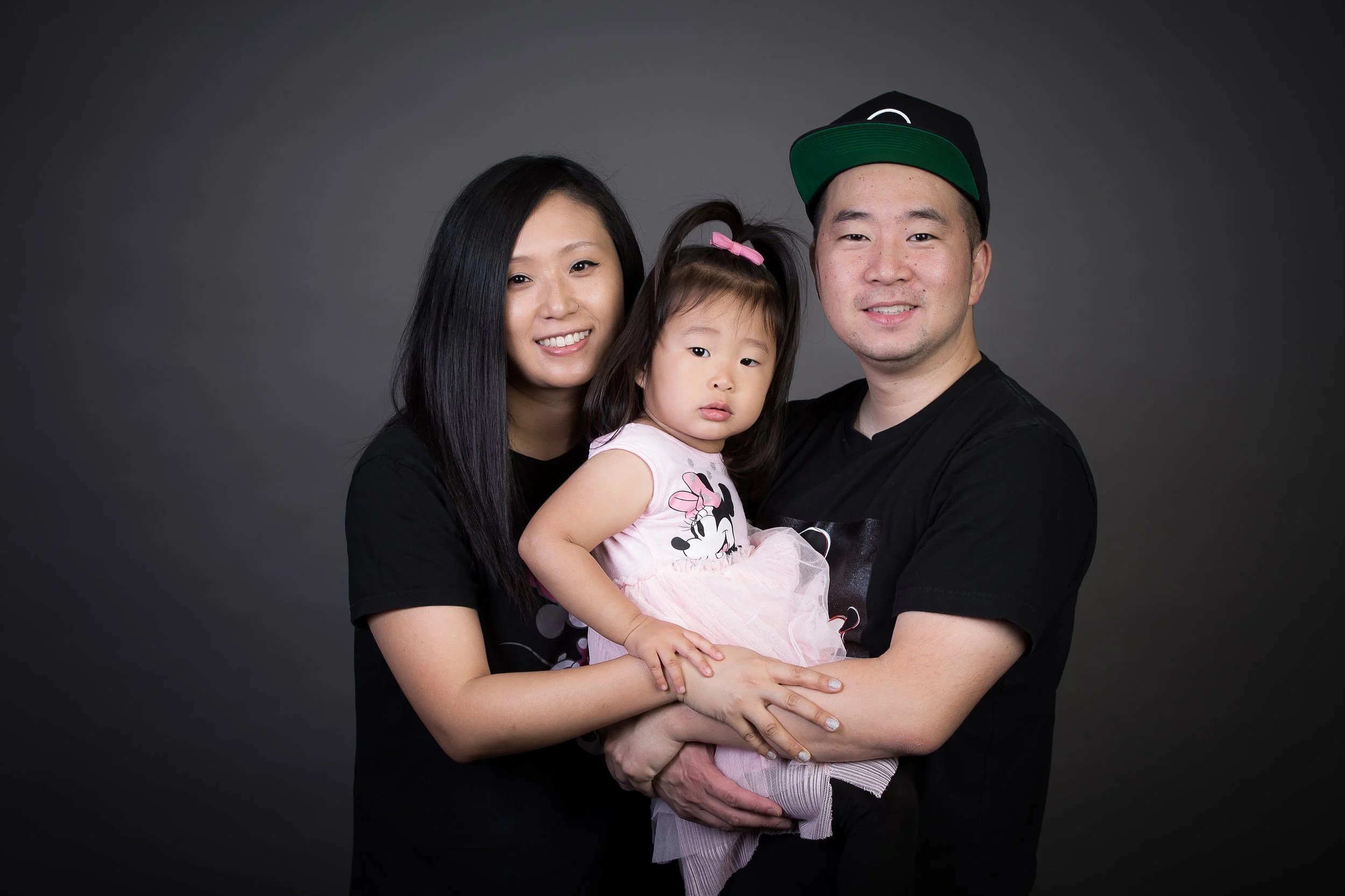 A family of three, including a woman, a man wearing a black cap, and a young girl, are posing together against a dark grey background. The woman and man are smiling, while the girl has a neutral expression and is wearing a pink dress with a Minnie Mo