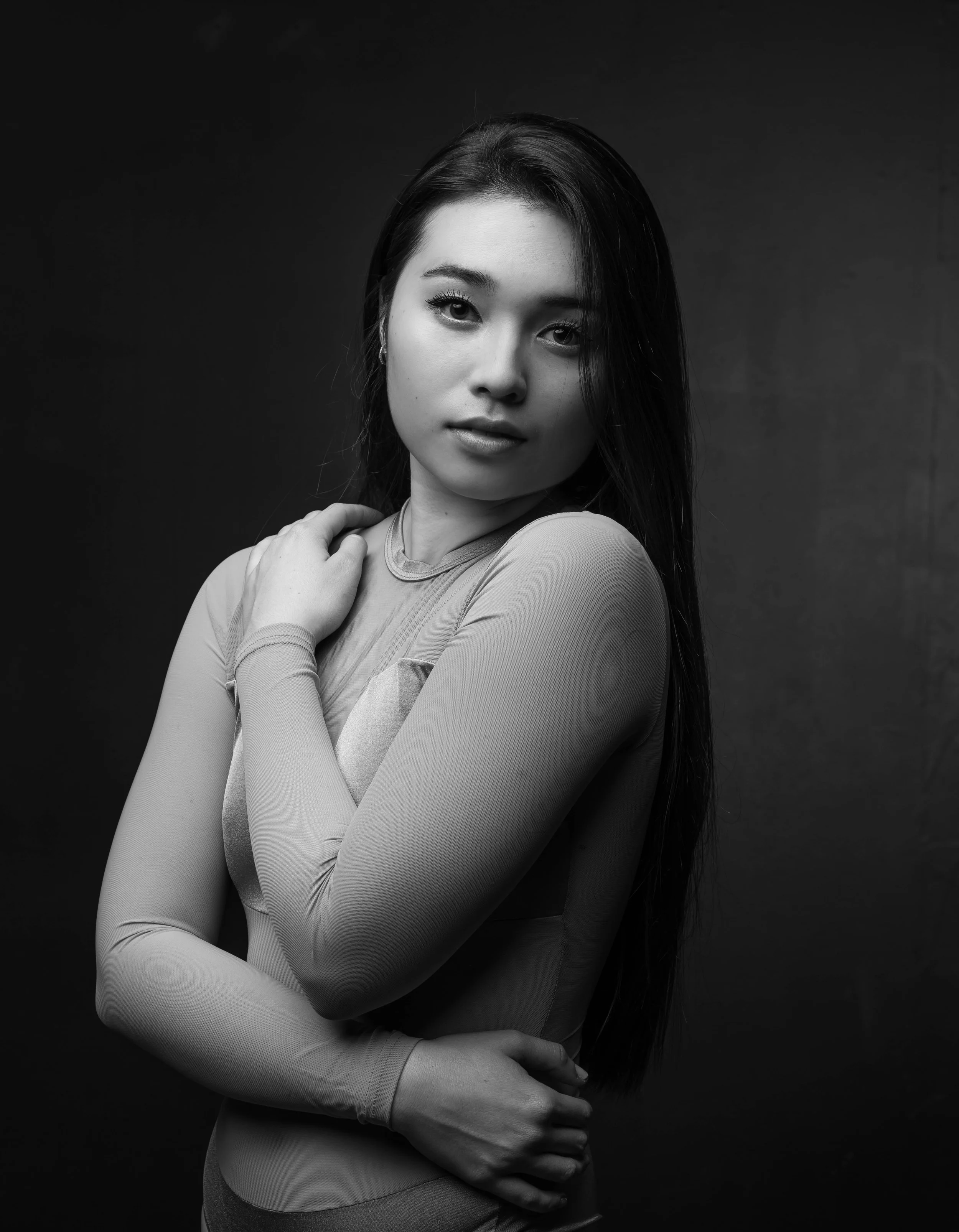 Black and white portrait of a woman with long hair, looking directly at the camera, with one hand near her neck and the other crossed over her waist, against a dark background.